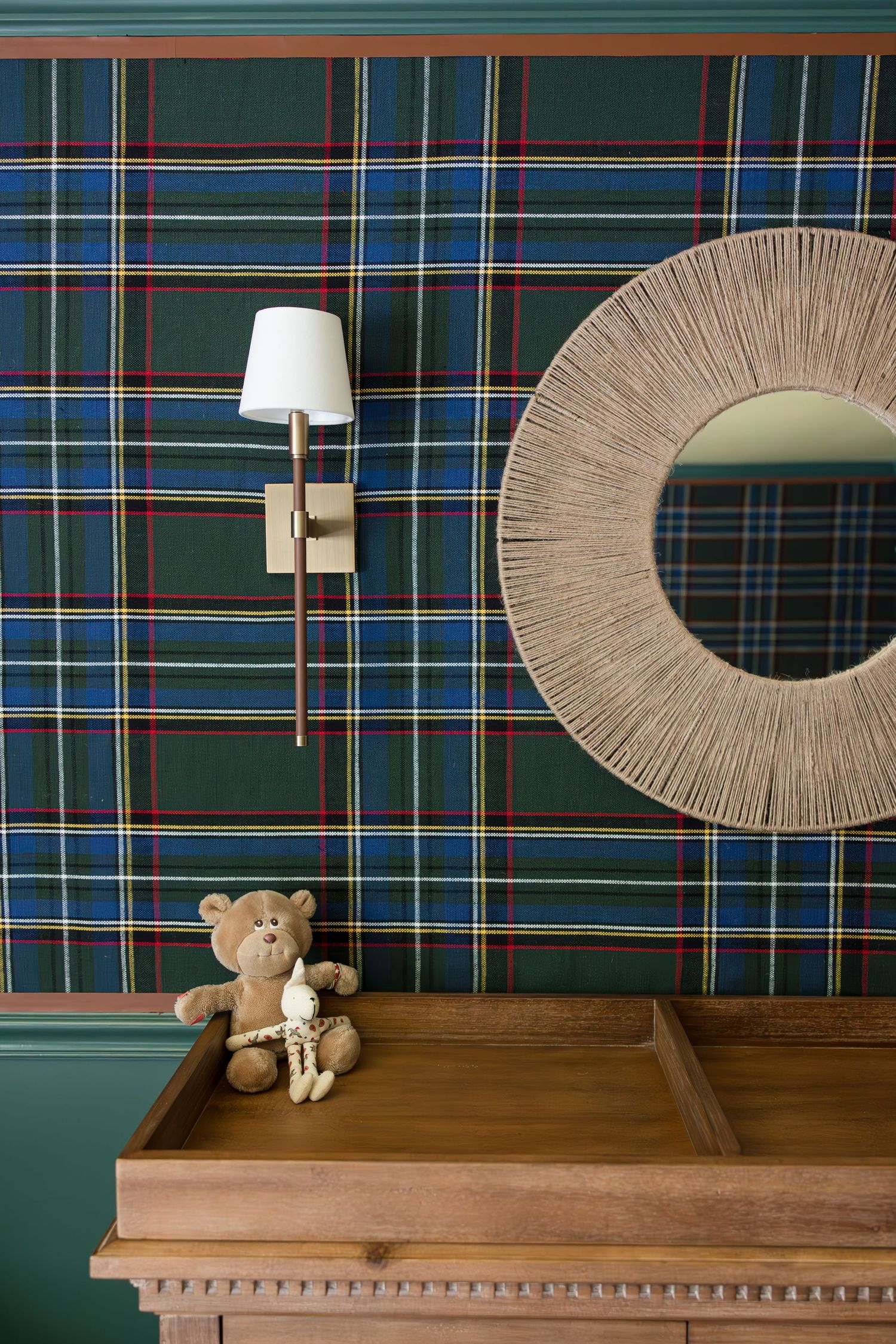 A wooden changing table with a small teddy bear beneath a circular woven mirror on plaid walls.