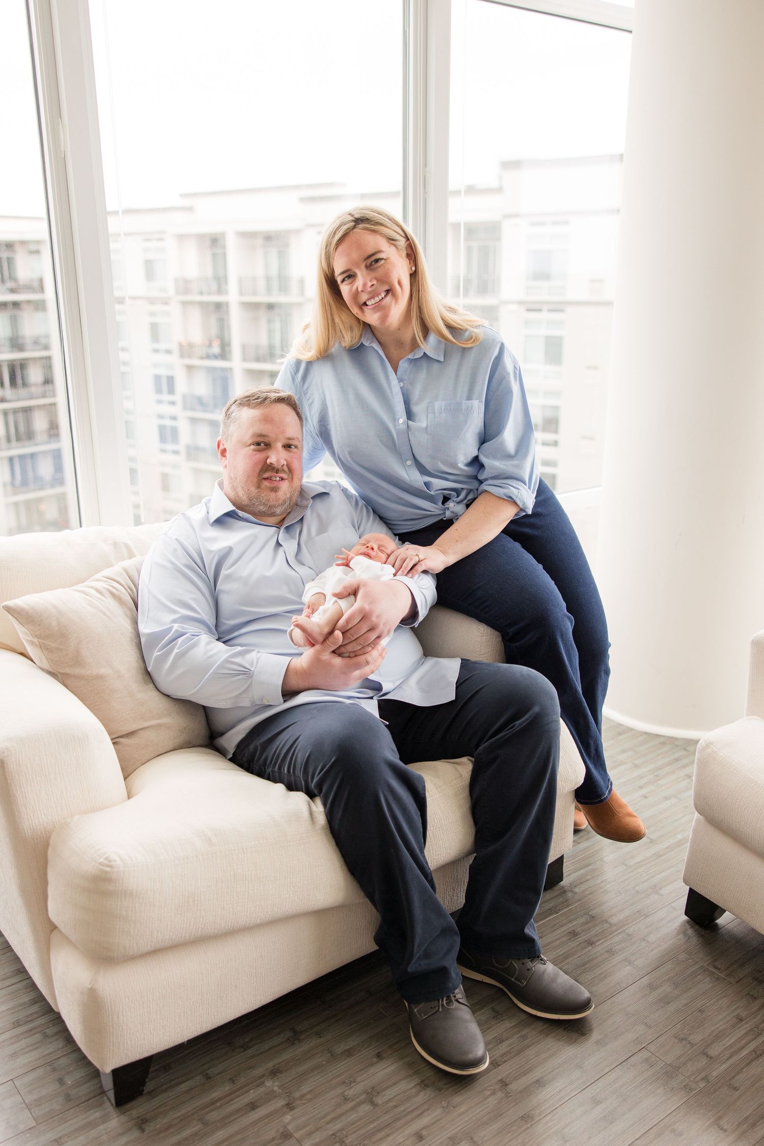 A couple relaxes together on a cream colored couch in a bright modern apartment with city views.