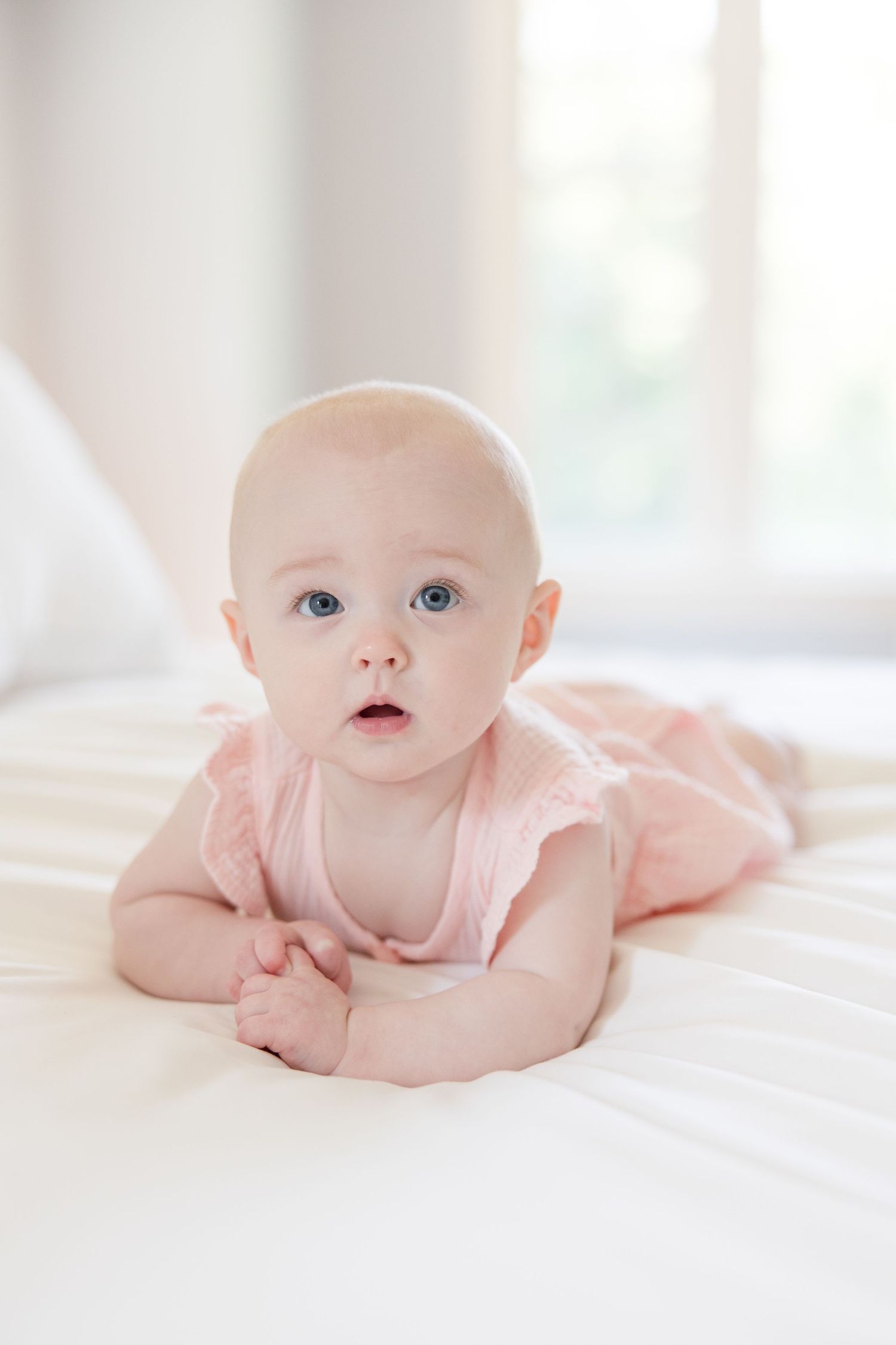 Beautiful baby in soft pink outfit lying on white bedding in natural light with a curious expression.
