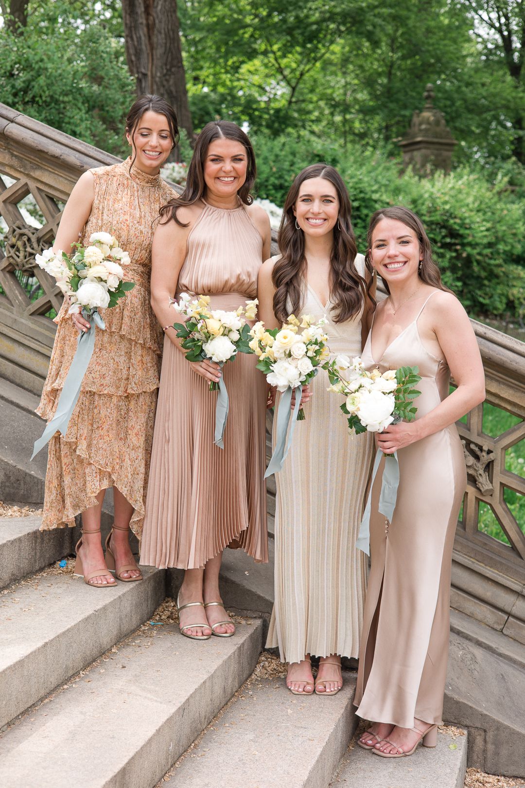 Four attendants in champagne and blush toned dresses hold white bouquets on stone steps.