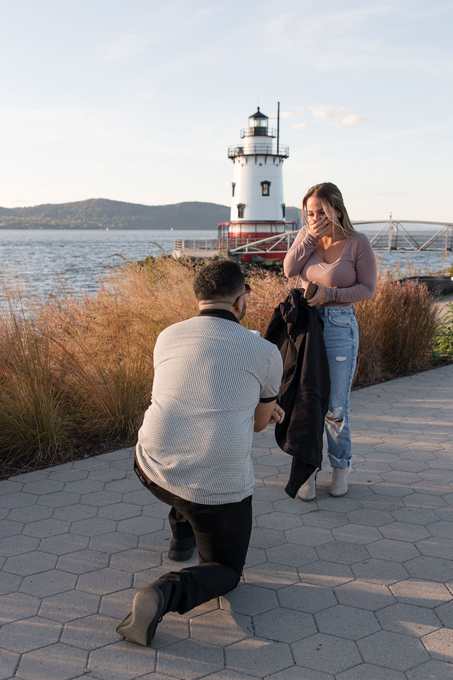Proposal scene at scenic lighthouse location with water and mountains in background.