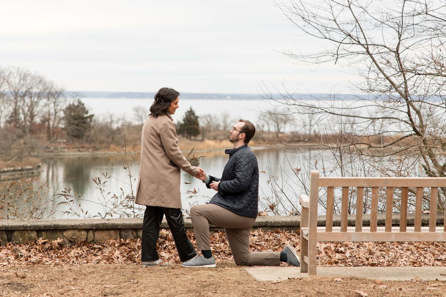 A romantic proposal moment captured on a park bench overlooking a serene lake on a winter day.