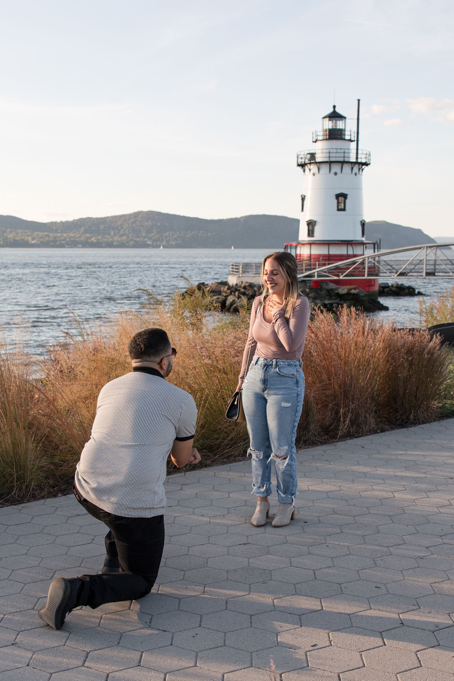 A proposal moment captured in front of a waterfront lighthouse with mountains in background.