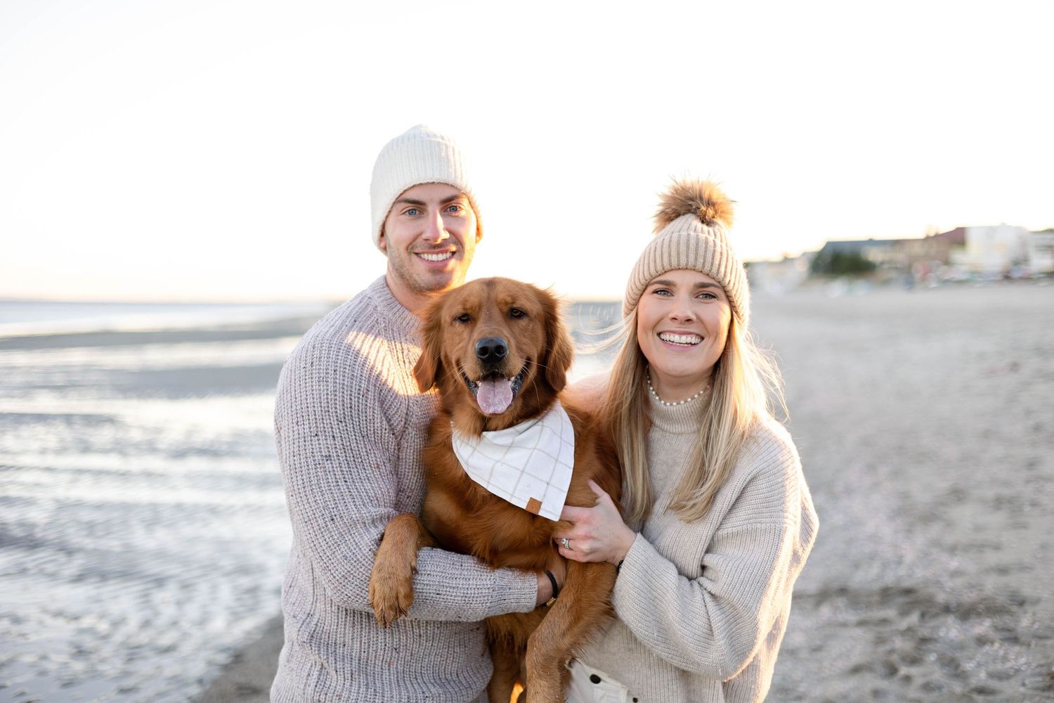 Happy couple holding golden retriever on beach in matching sweaters.