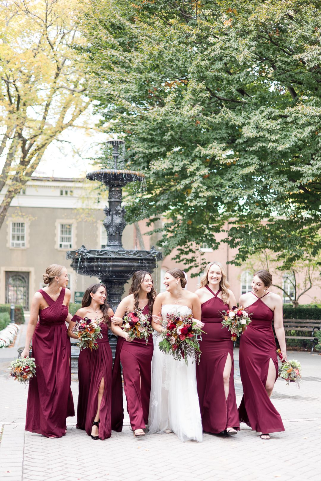 A bridal party in burgundy dresses poses around a fountain in an outdoor garden setting.