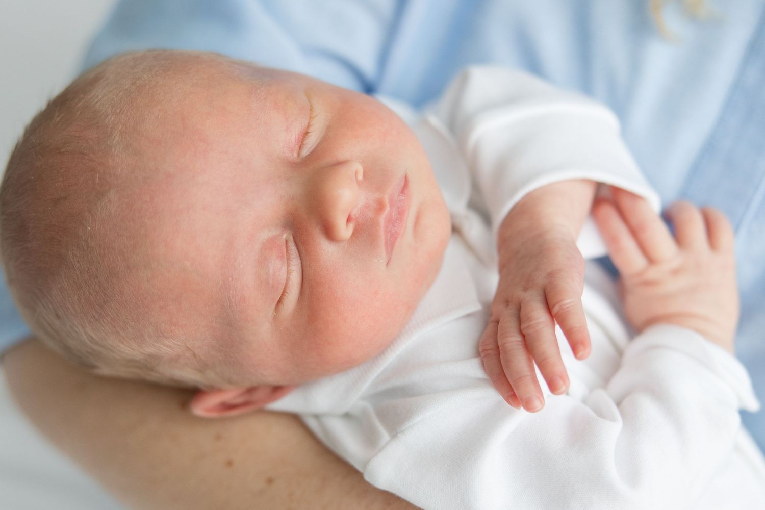 A peaceful sleeping newborn baby wearing white clothing rests on blue bedding.
