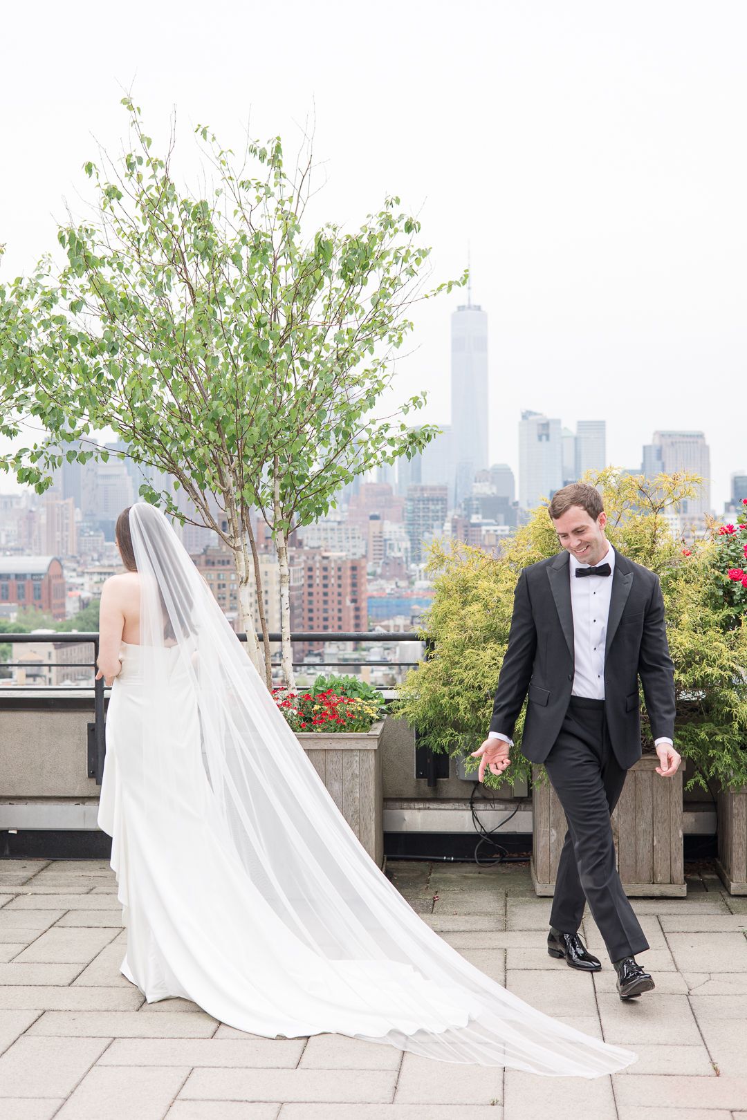 Wedding couple walking separately on a rooftop terrace with flowing veil and NYC skyline backdrop.