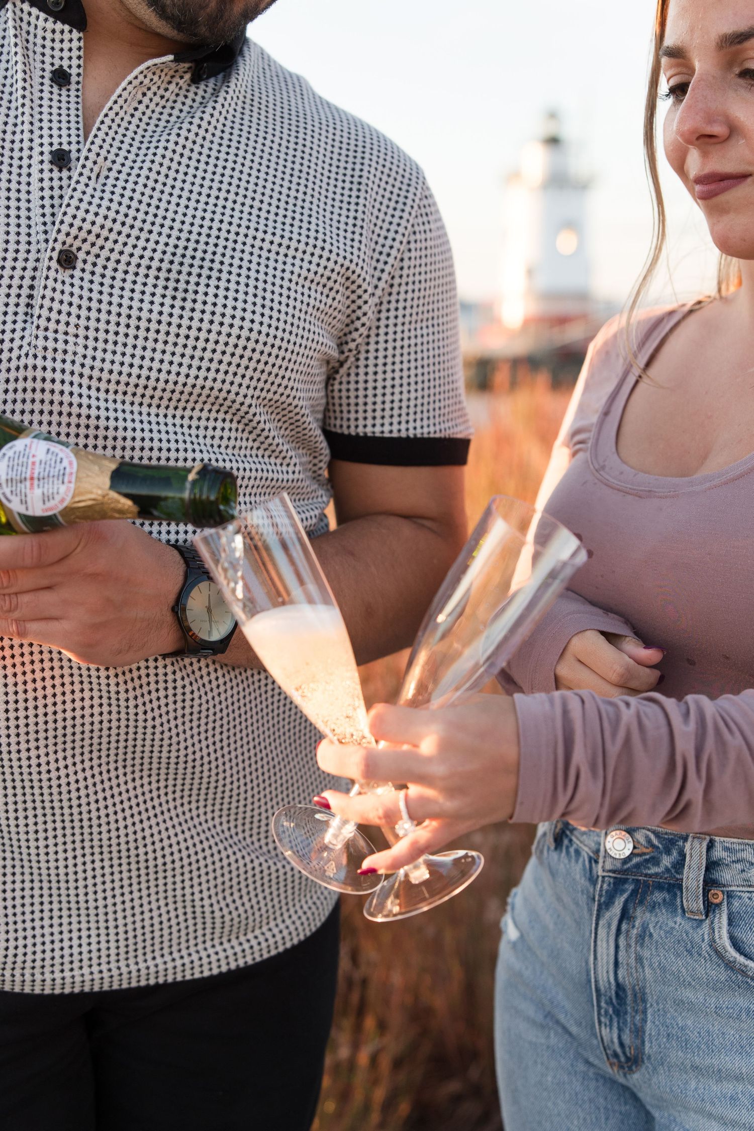 Close-up of hands holding champagne glasses against a sunset lighthouse backdrop.