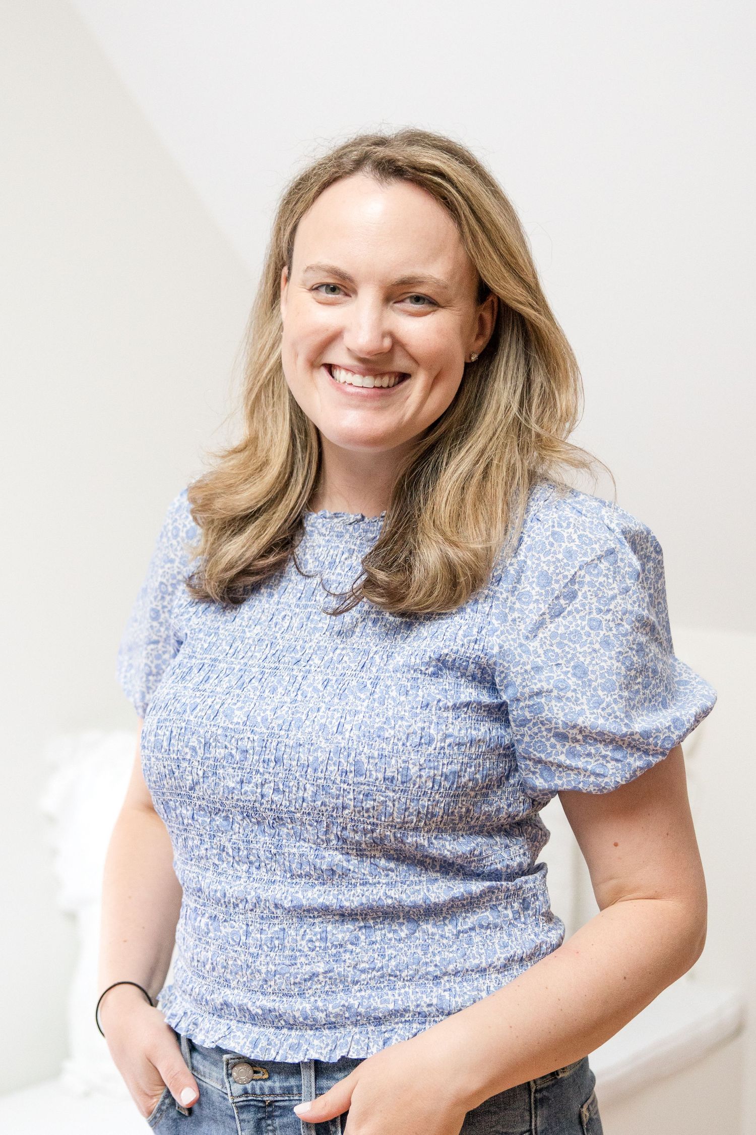 A person wearing a light blue puff sleeve top and jeans smiles warmly against a white background.