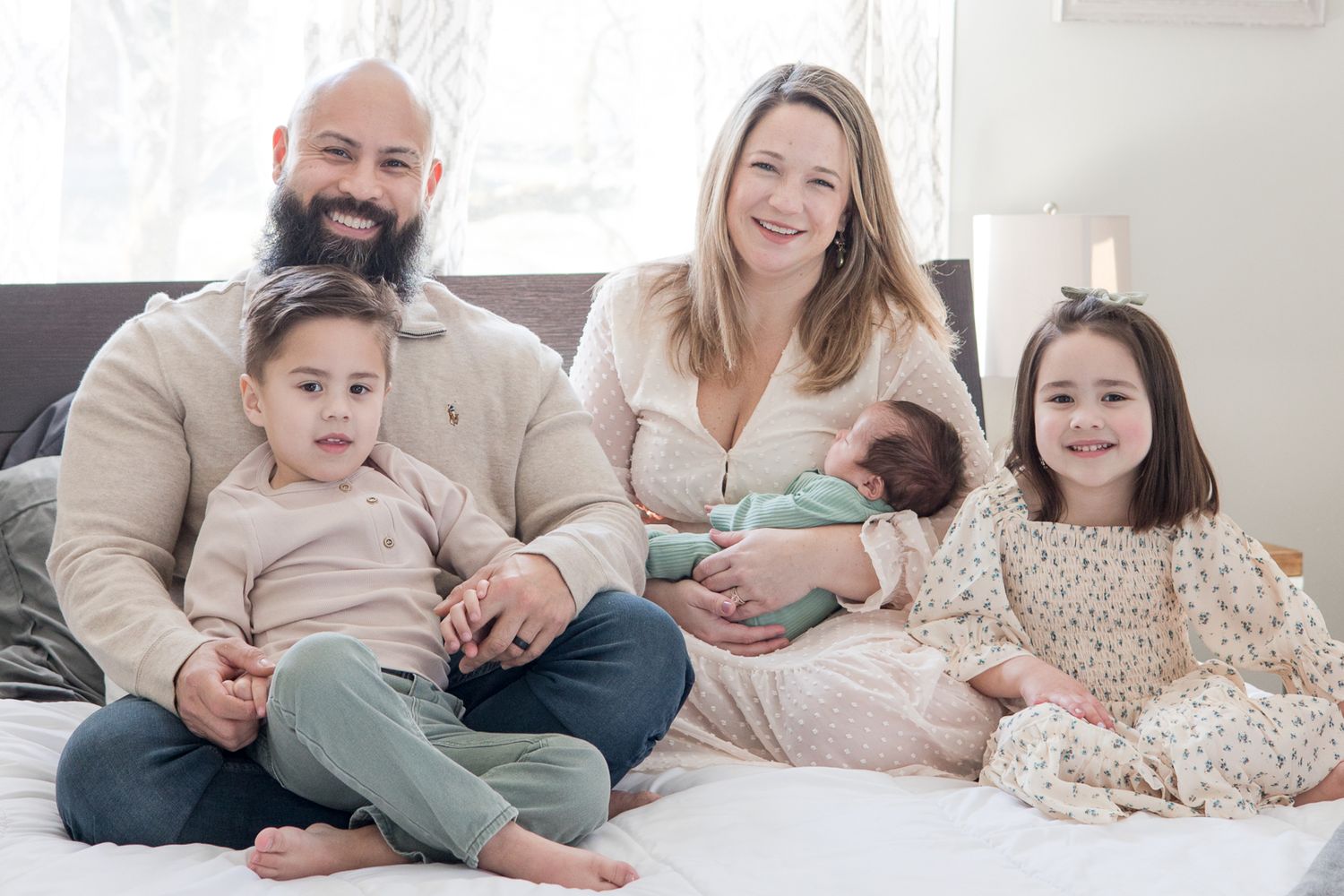 A family relaxes together on a bed wearing coordinated casual outfits in a light-filled bedroom.