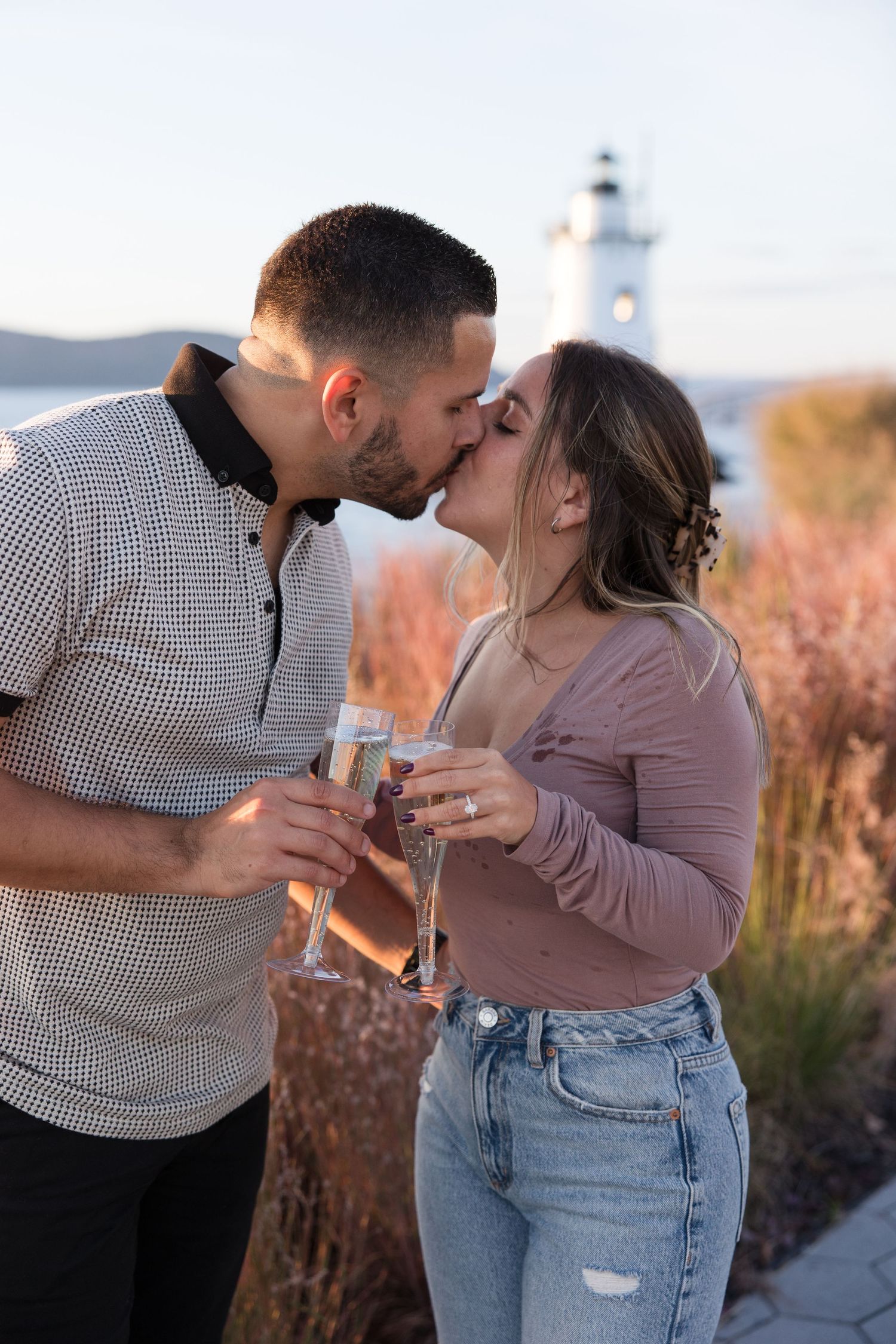 A couple shares intimate moment with champagne glasses by lighthouse at sunset.