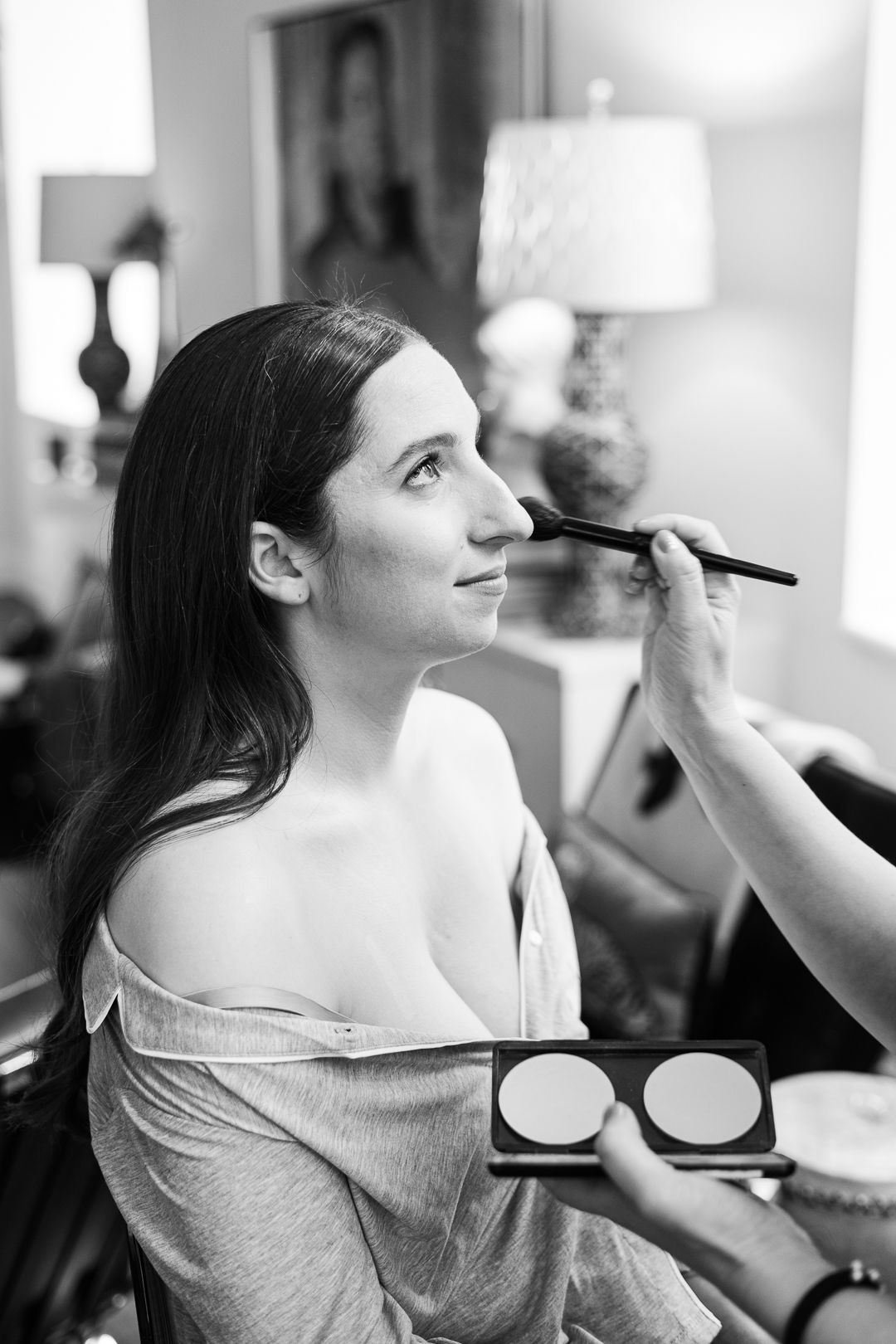 Black and white photograph of bridal makeup application in intimate setting.