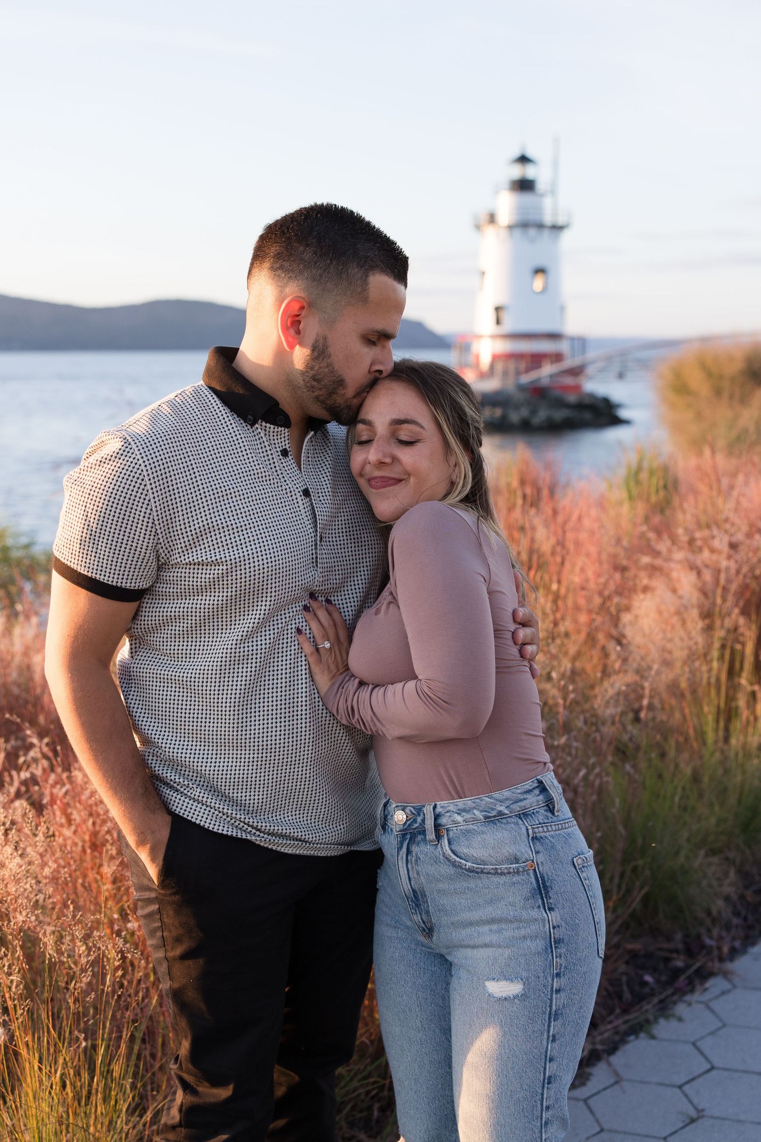 Couple sharing intimate moment near coastal lighthouse during golden hour.
