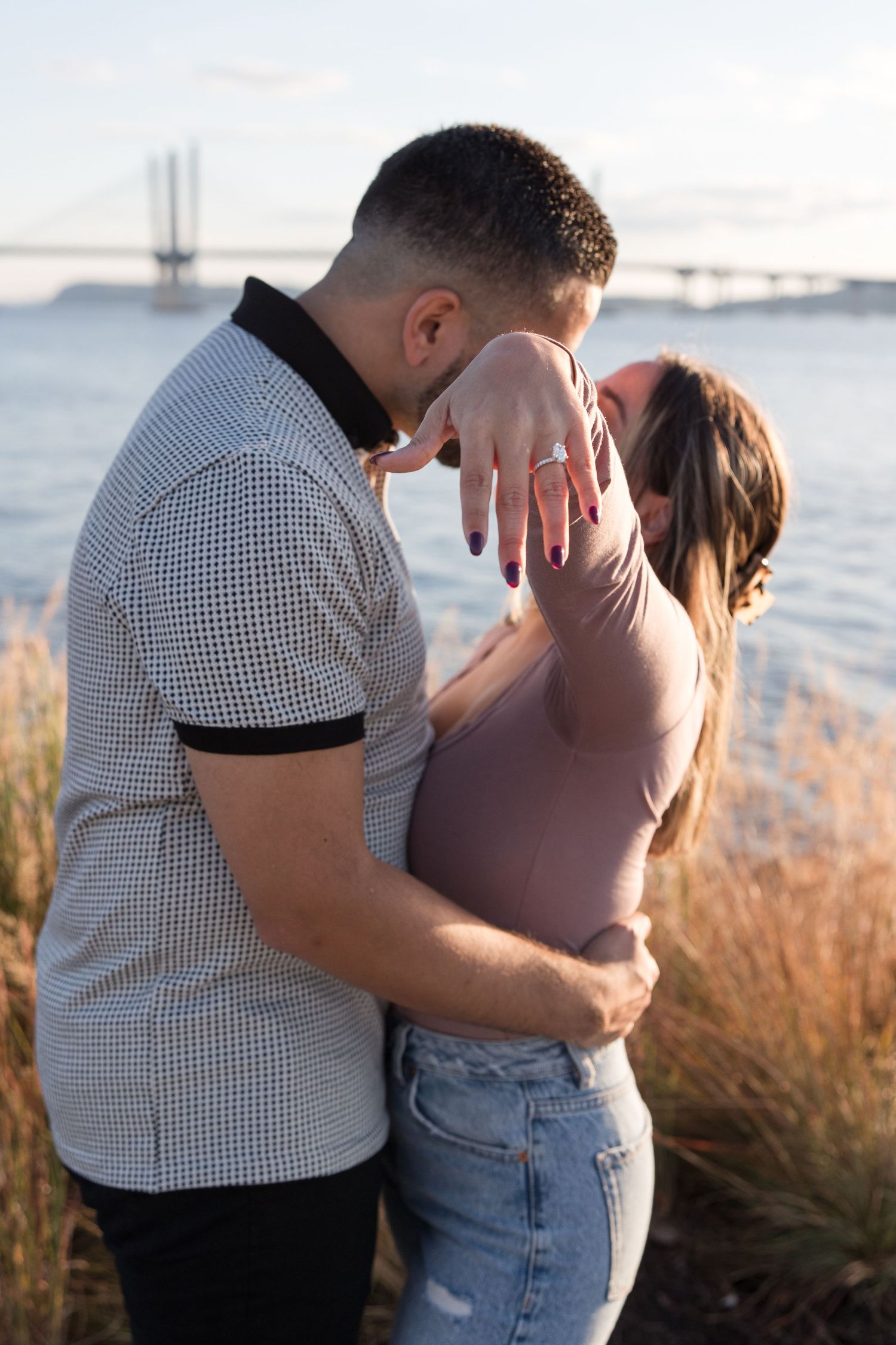 Newly engaged couple embracing by the water with bridge in distance.