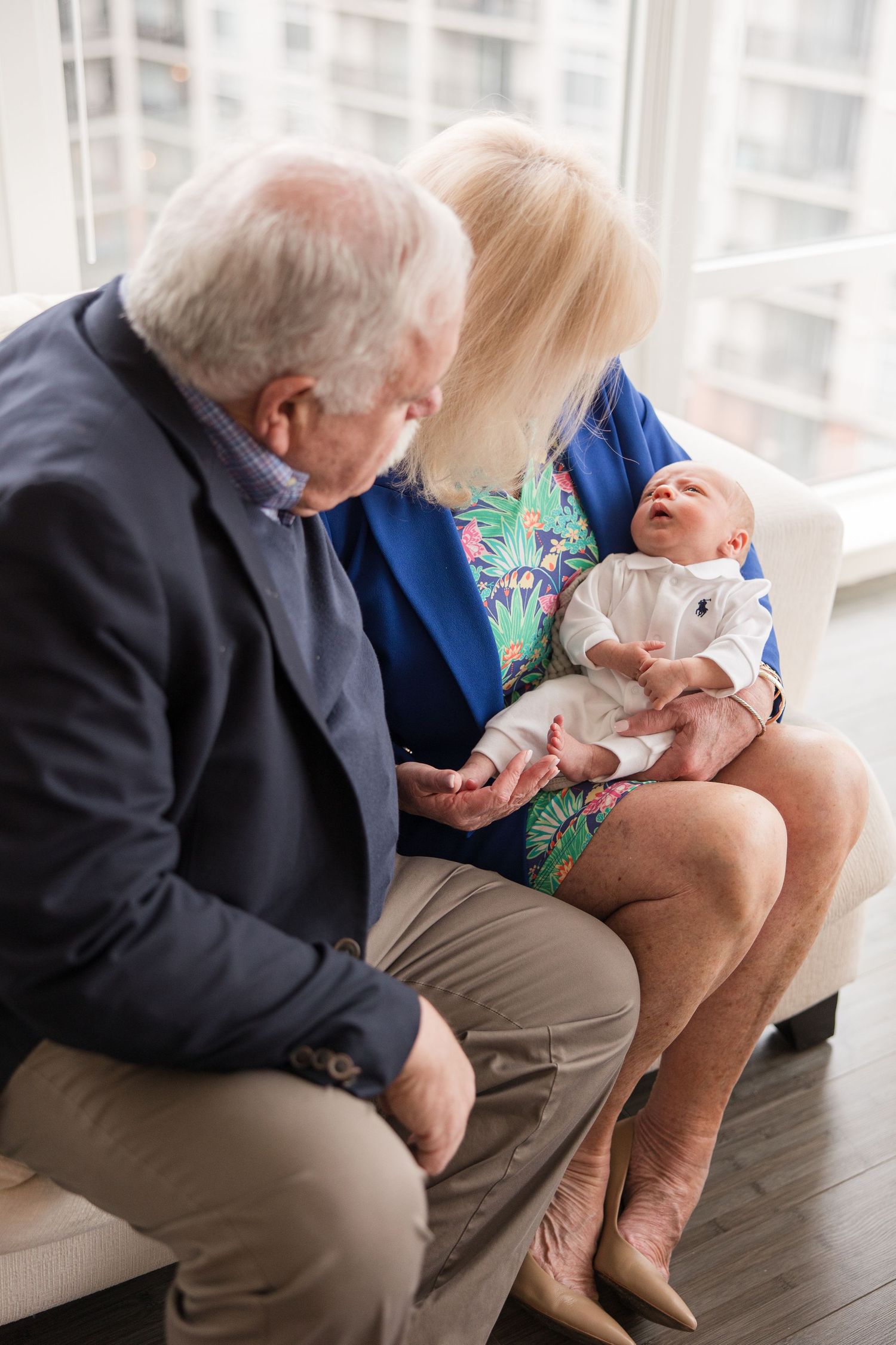 An elderly couple sitting together on a couch holds and admires a tiny newborn baby dressed in white.