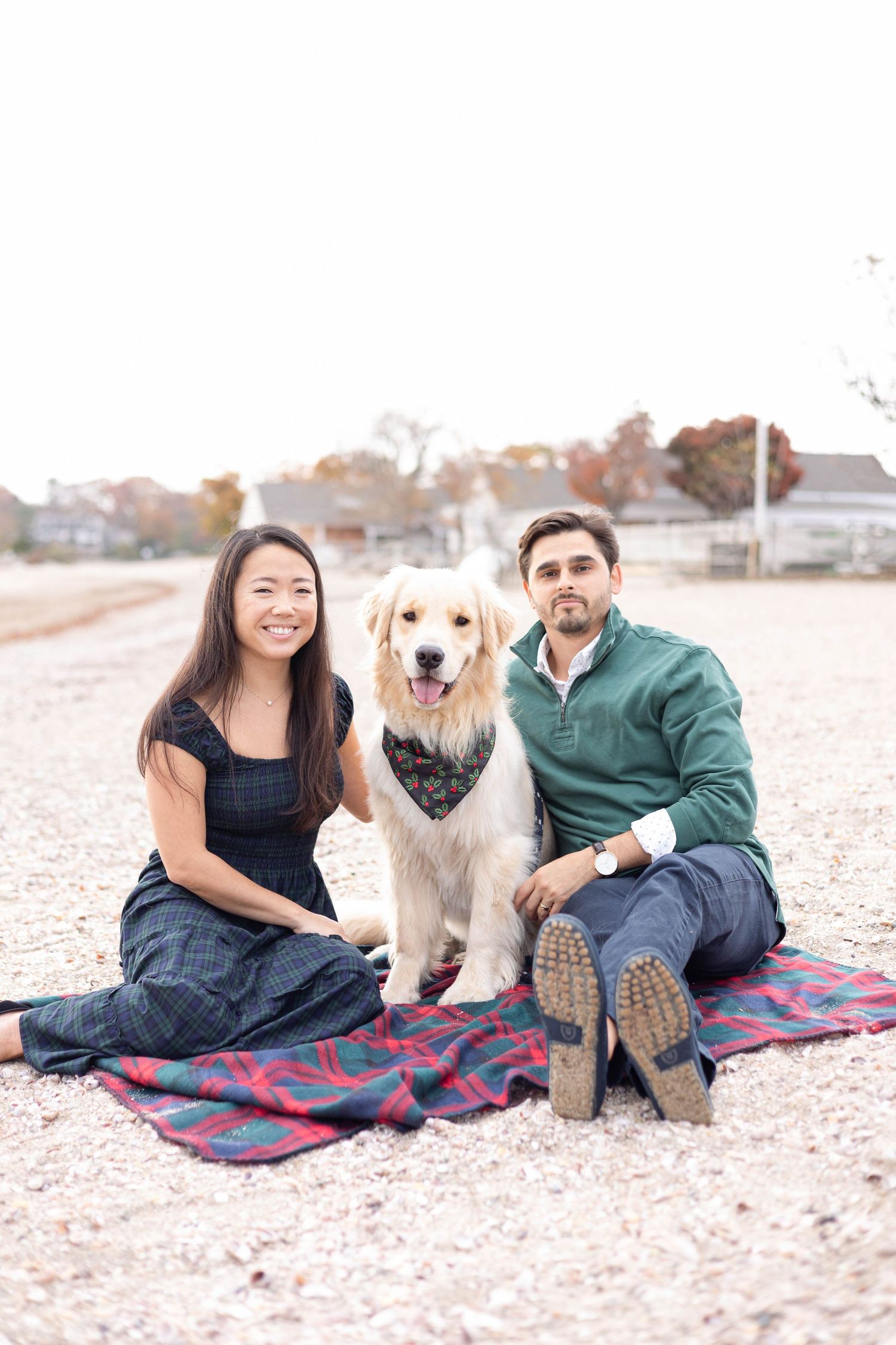 A couple sits with their happy golden retriever on a plaid blanket at the beach.