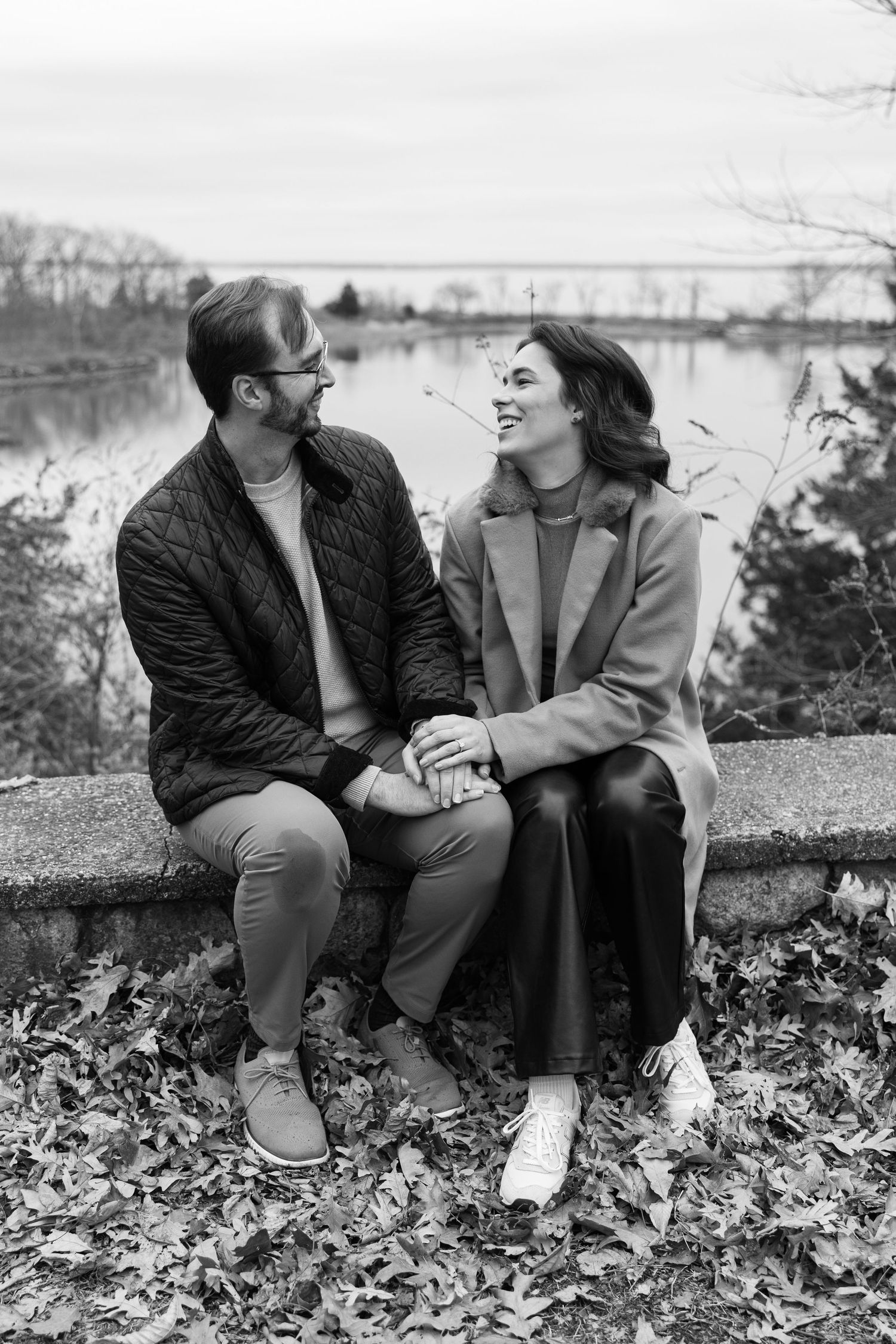A couple sitting together and laughing on a stone wall surrounded by autumn leaves.