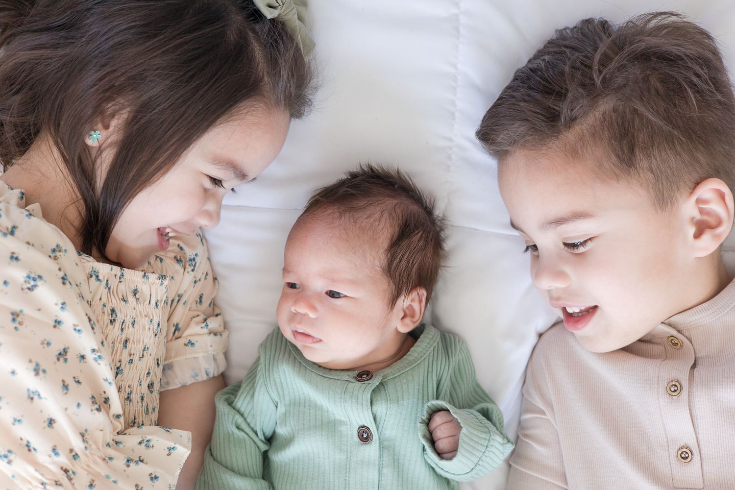 Three siblings lay close together on white bedding wearing coordinated outfits.