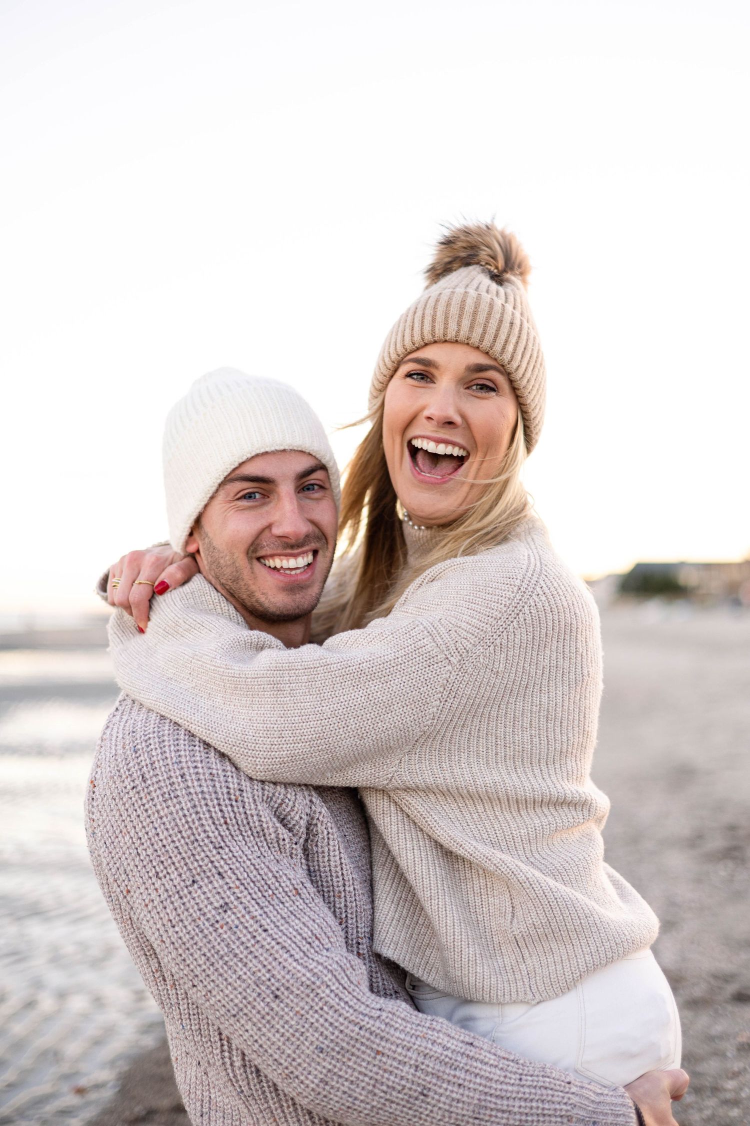 Couple in knit sweaters and beanies laughing on beach.