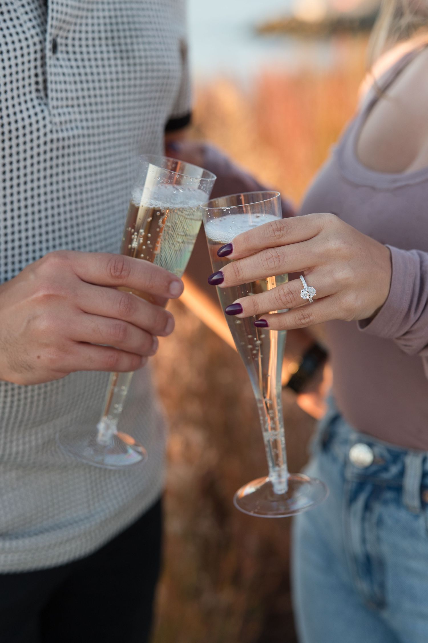 Two hands clink champagne glasses together in celebration with engagement ring visible.