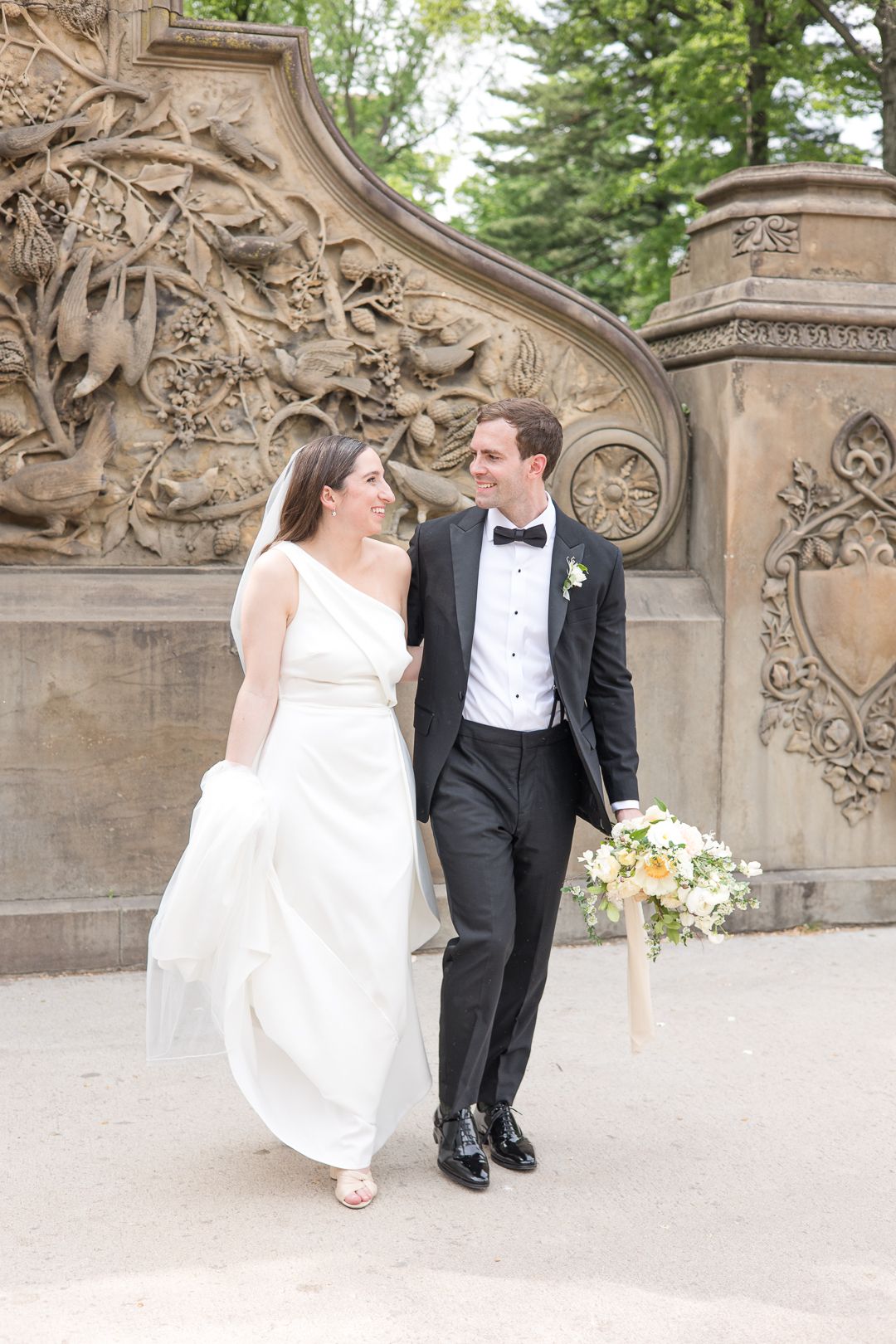Newlyweds share intimate moment while walking together against decorative stone wall background.