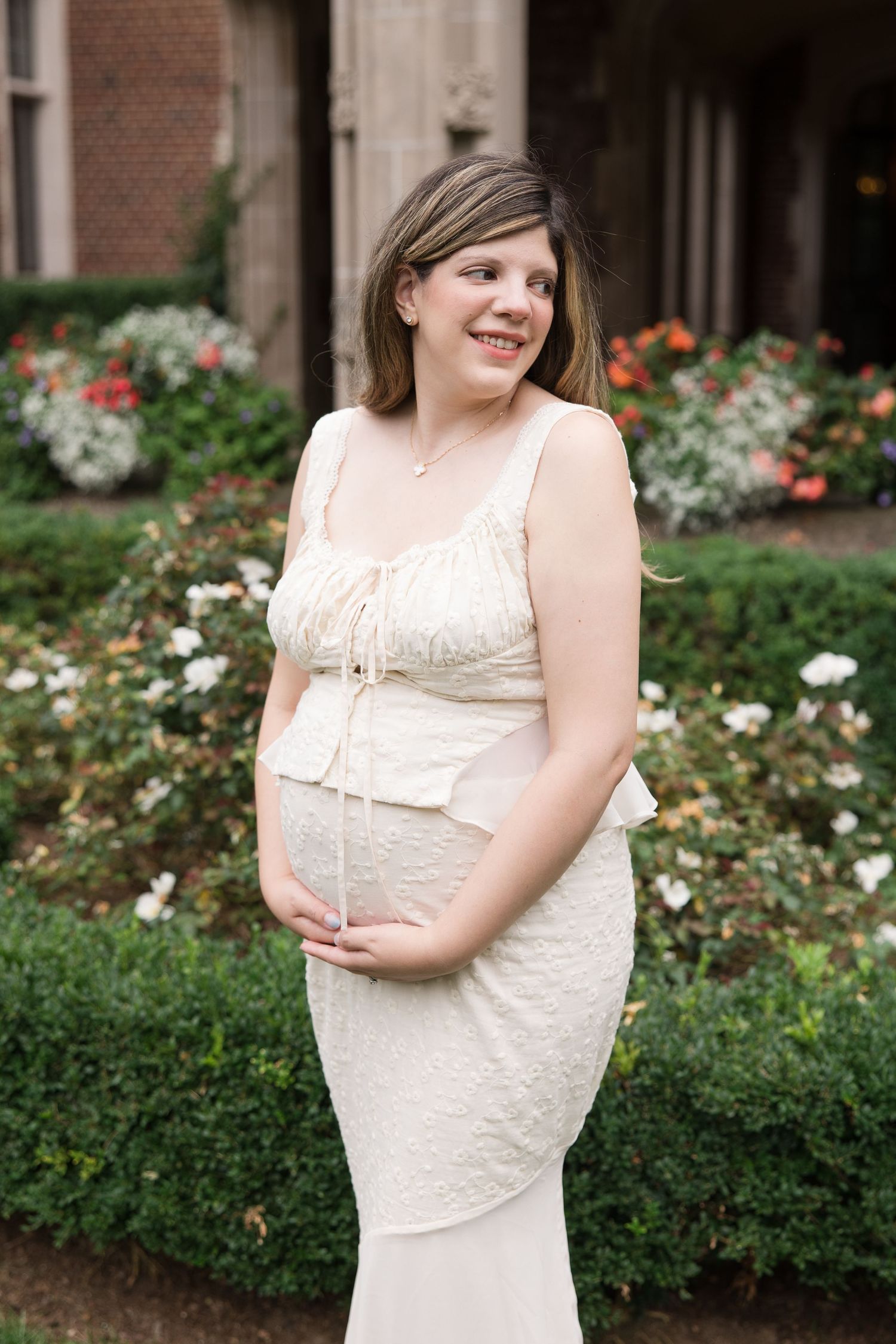 A person in a sleeveless cream lace dress stands in front of a floral garden with roses and brick building backdrop.