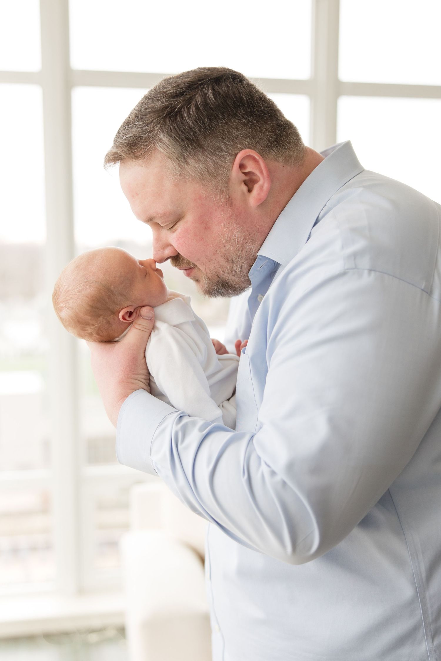 A tender moment between a parent in a light blue button-down shirt and a newborn baby near a bright window.