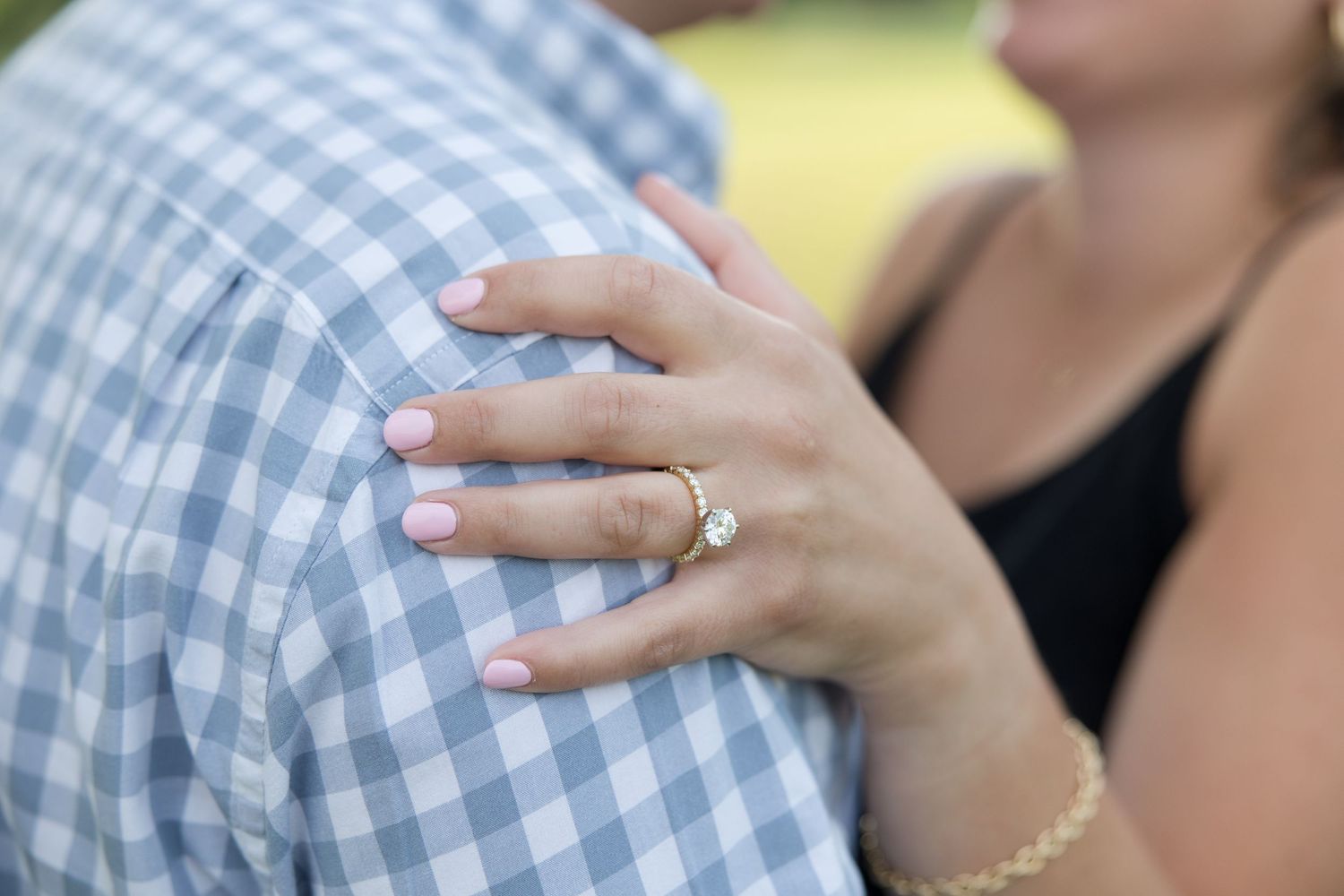 A delicate engagement ring sparkles as hands rest against a blue and white checked shirt pattern.