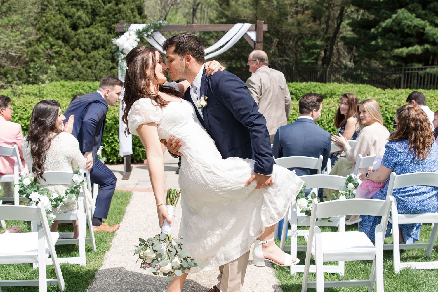 Romantic outdoor wedding kiss captured in a garden setting with white chairs.