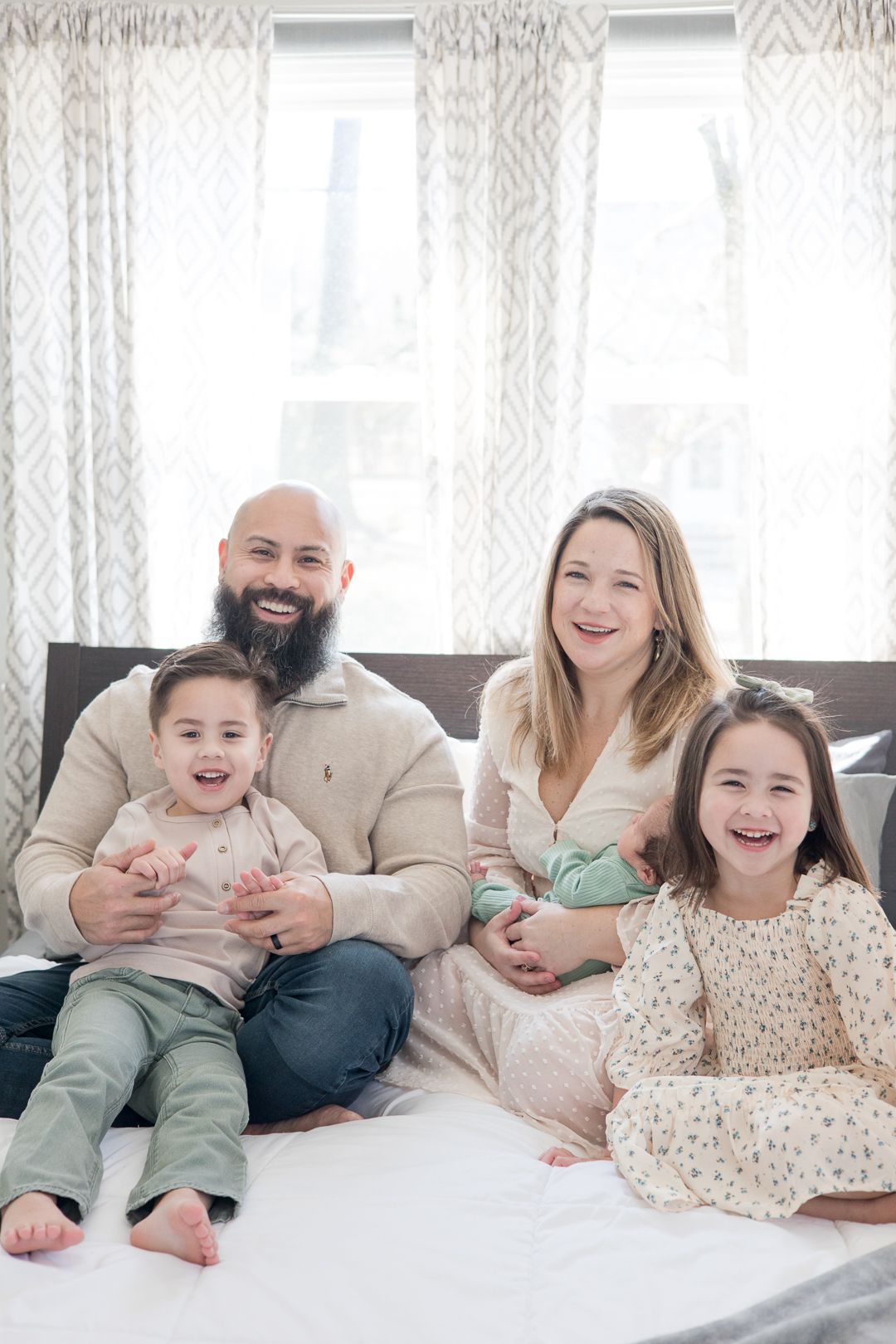 Family relaxing together on bed with patterned curtains and natural light.