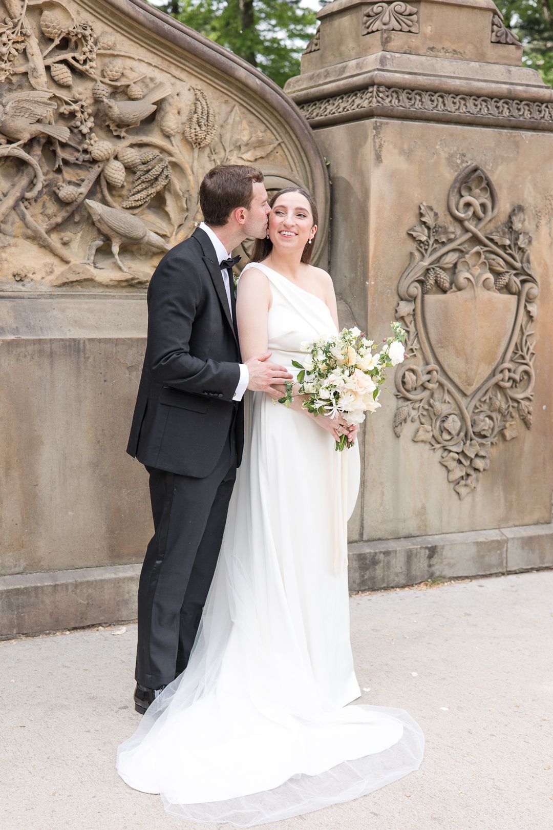 Elegant wedding couple embraces by ornate stone wall backdrop in formal attire.