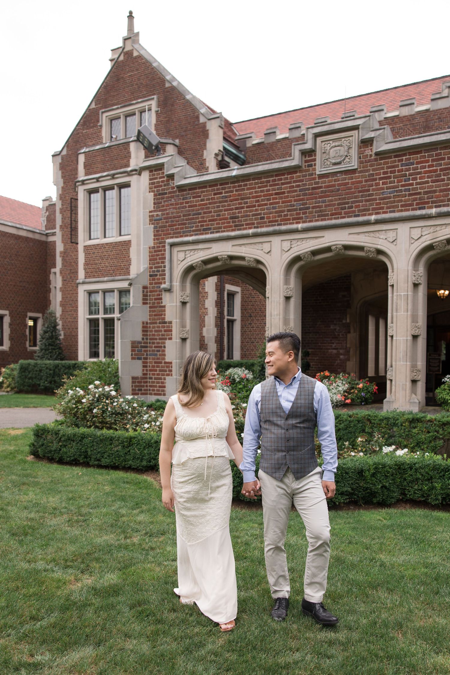 Couple poses together on lawn in front of historic brick building with arched doorways.