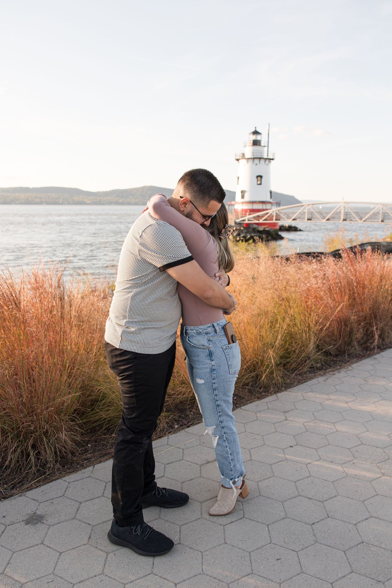A couple embraces near a lighthouse during sunset with water and mountains in background.