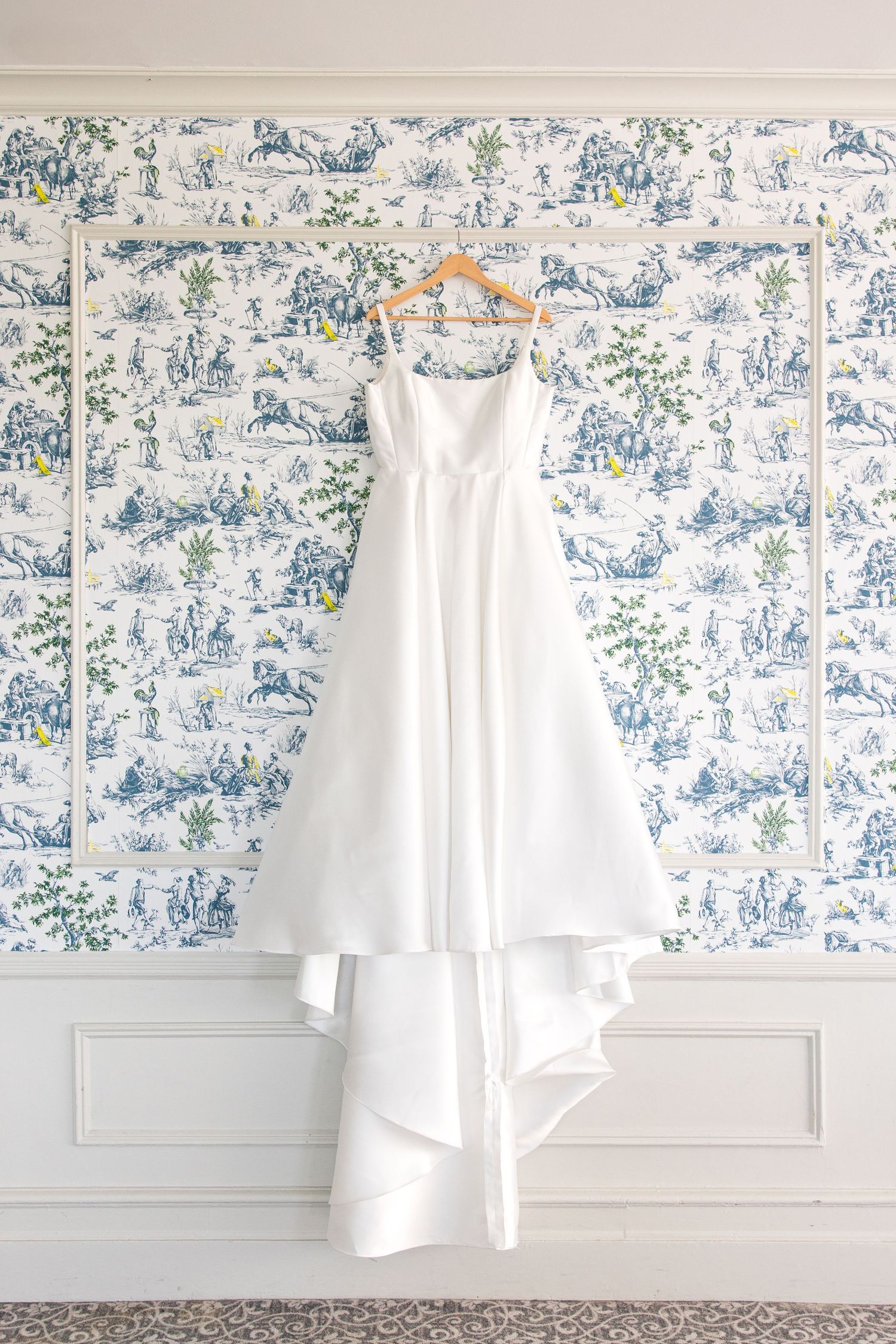 A classic white wedding dress hangs on a wooden hanger against blue and white toile wallpaper in an elegant room.