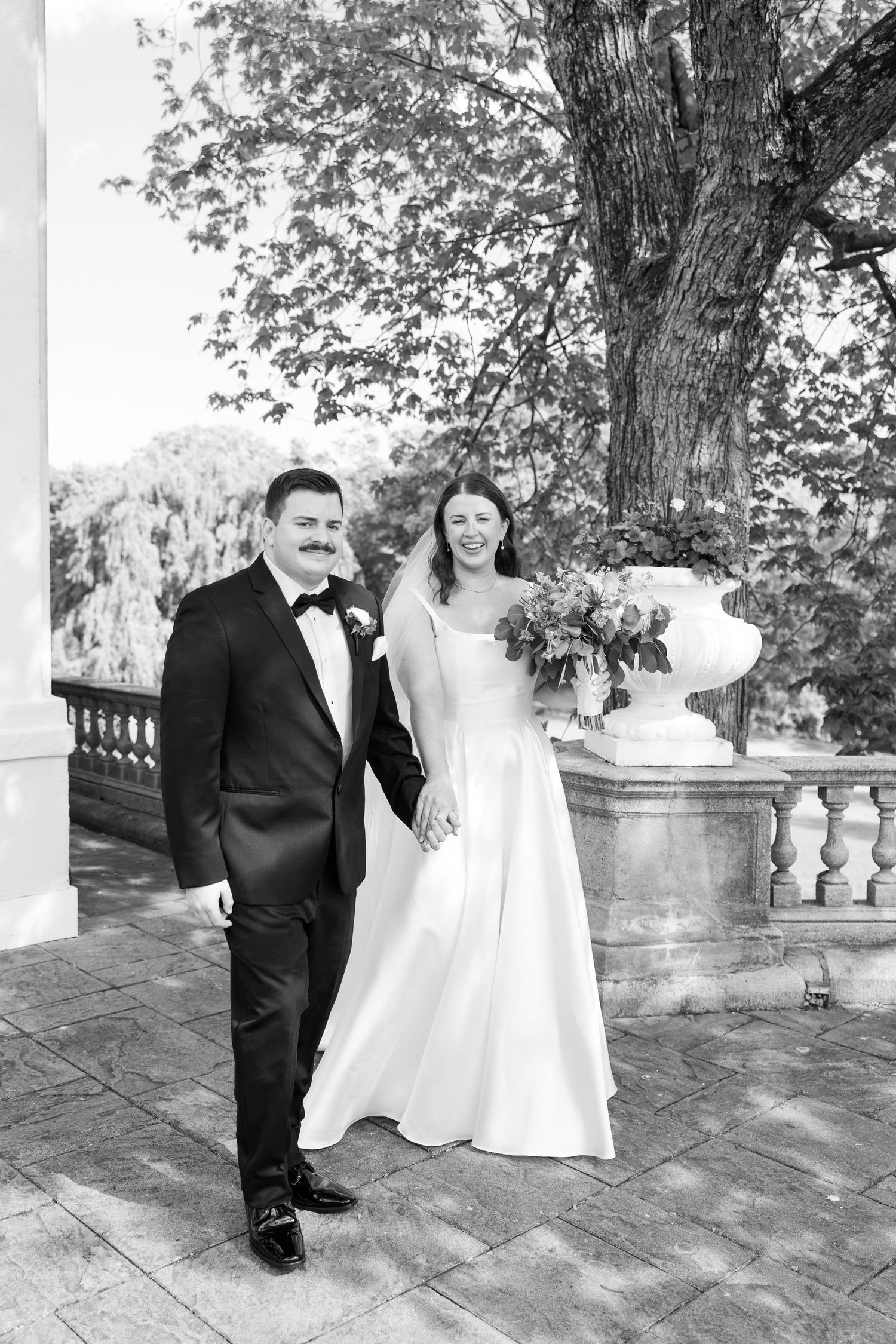A black and white wedding photo of a couple standing on a stone terrace beside a large tree and decorative stone railing.