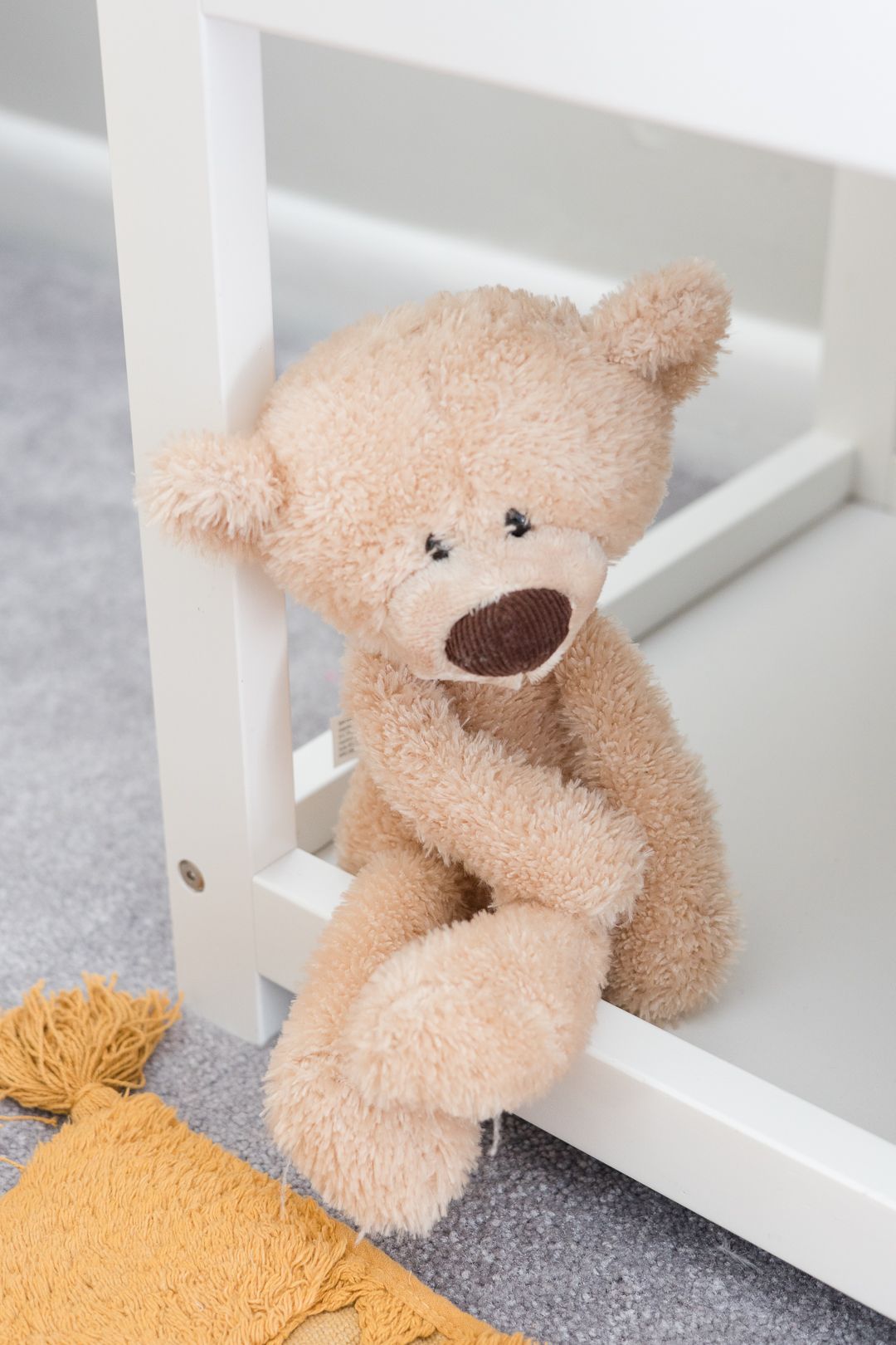 Plush beige teddy bear sitting against white furniture on gray carpet.