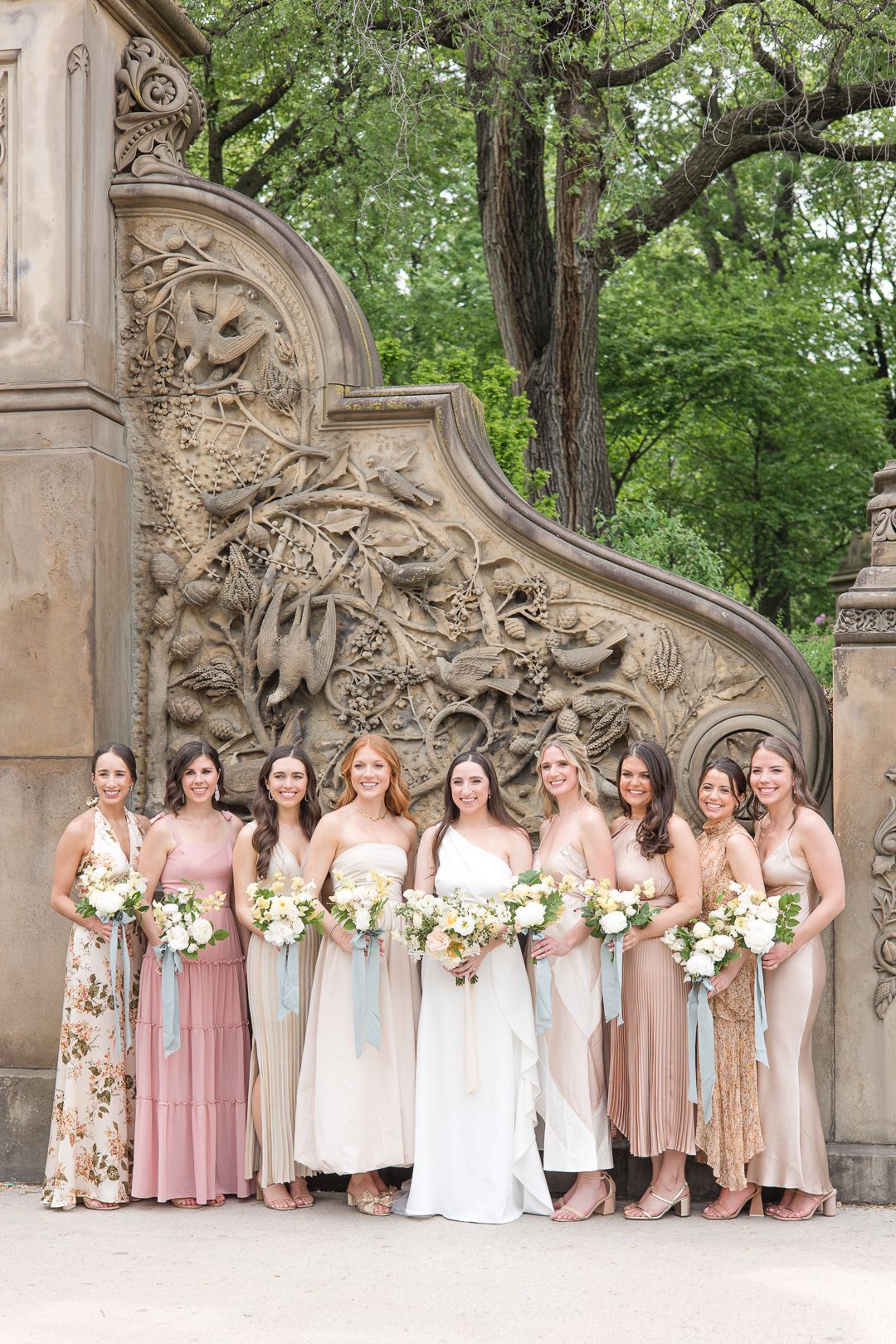 Bridal party in soft pink and neutral dresses posed against ornate stone wall.