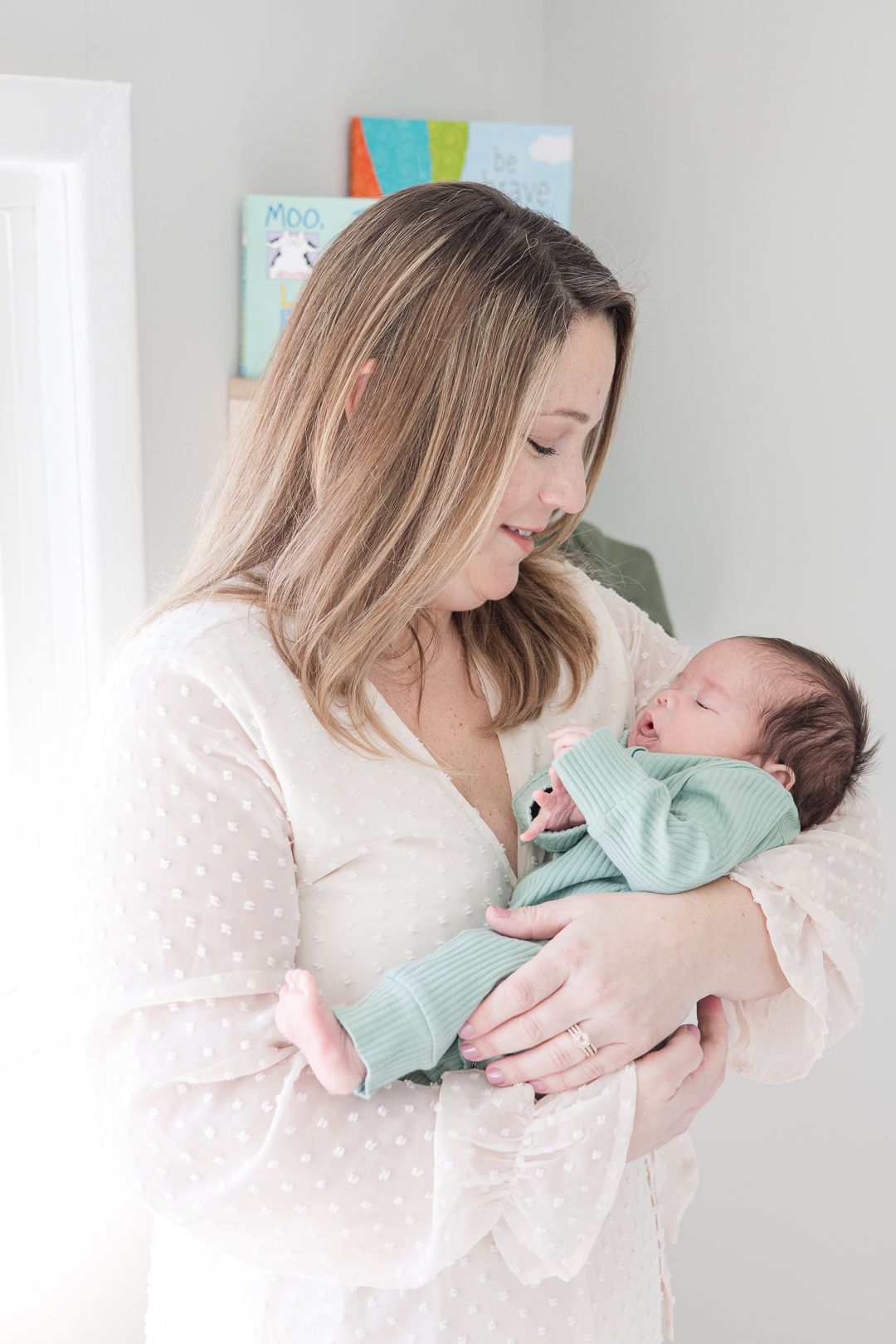 A woman in a polka dot dress holds a newborn baby wearing mint green against a white wall with artwork.