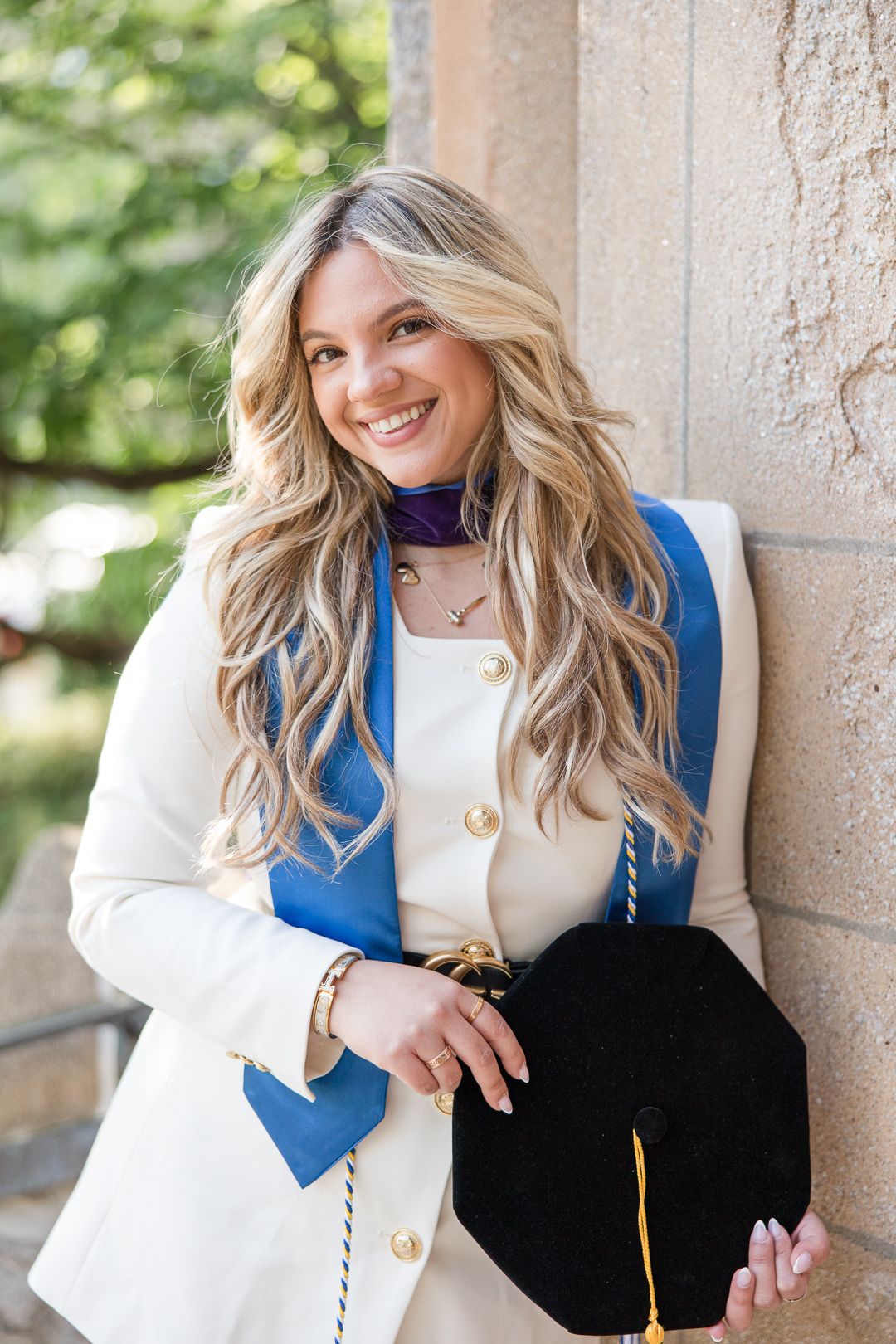 Graduation portrait against stone column wearing white blazer and blue vest.