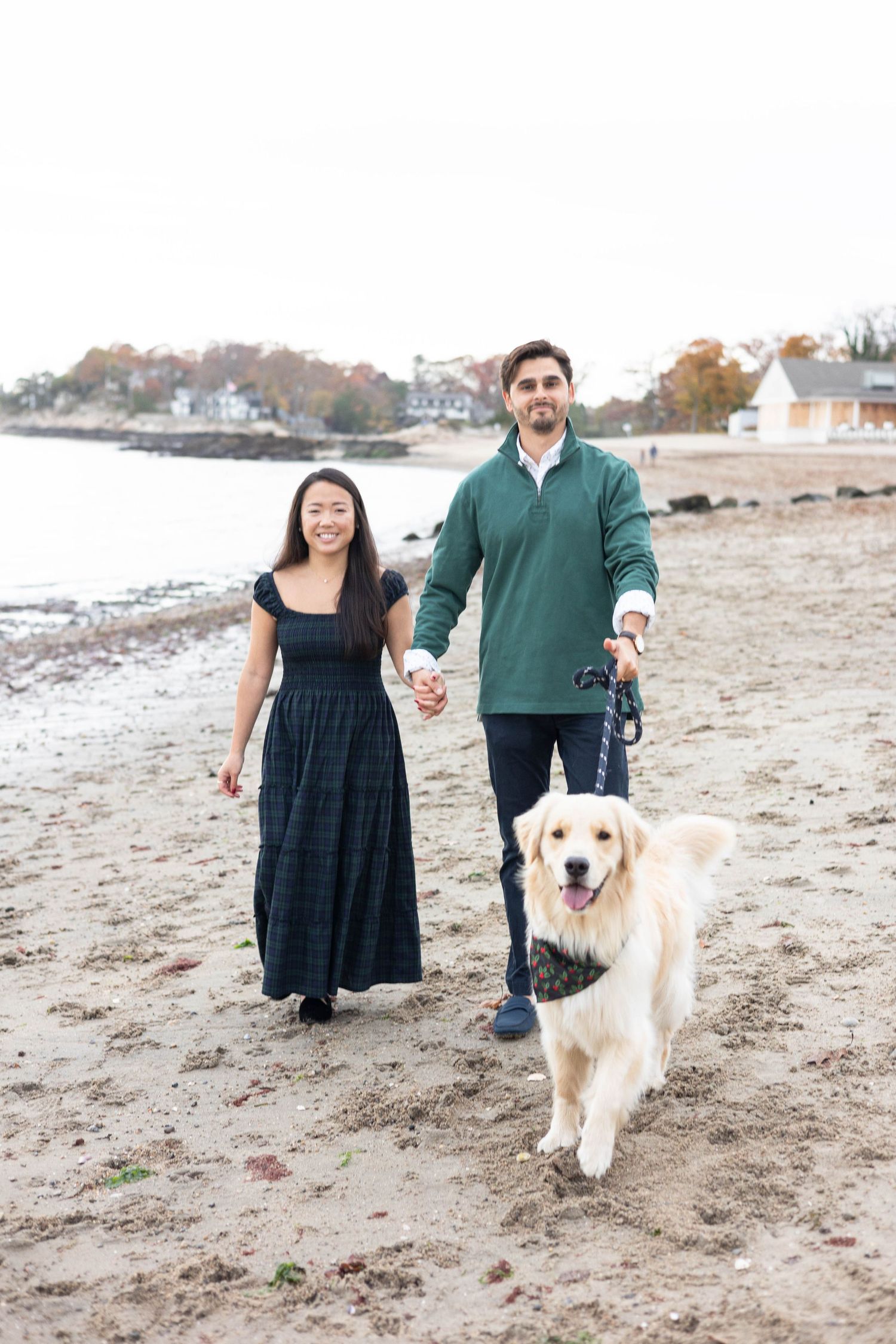Couple walks their Golden Retriever along sandy beach on cloudy day.