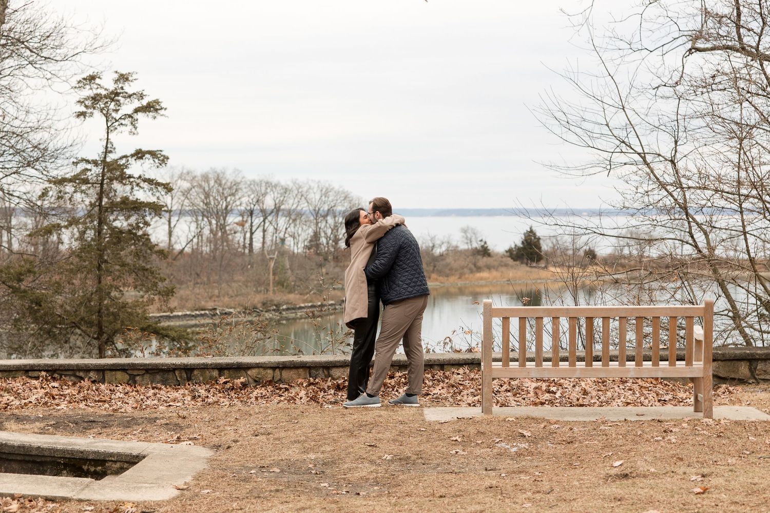 A couple embracing on a leaf-covered path overlooking a scenic lake view.