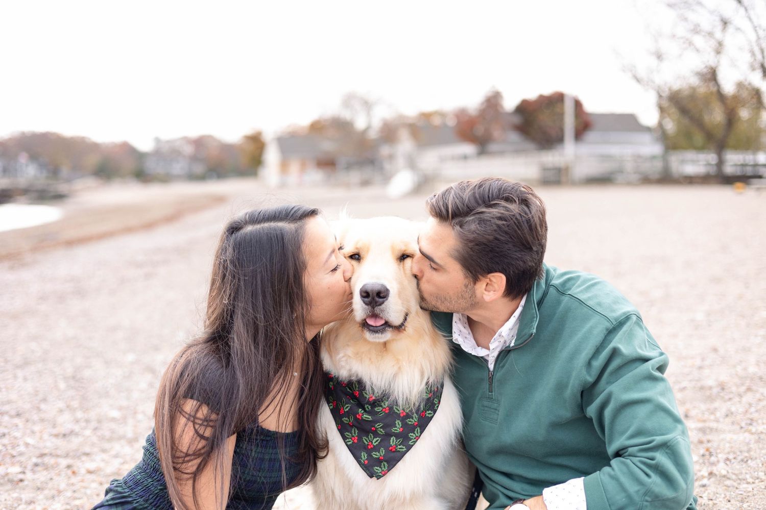 A couple shares an intimate moment with their smiling golden retriever wearing a bandana on the beach.