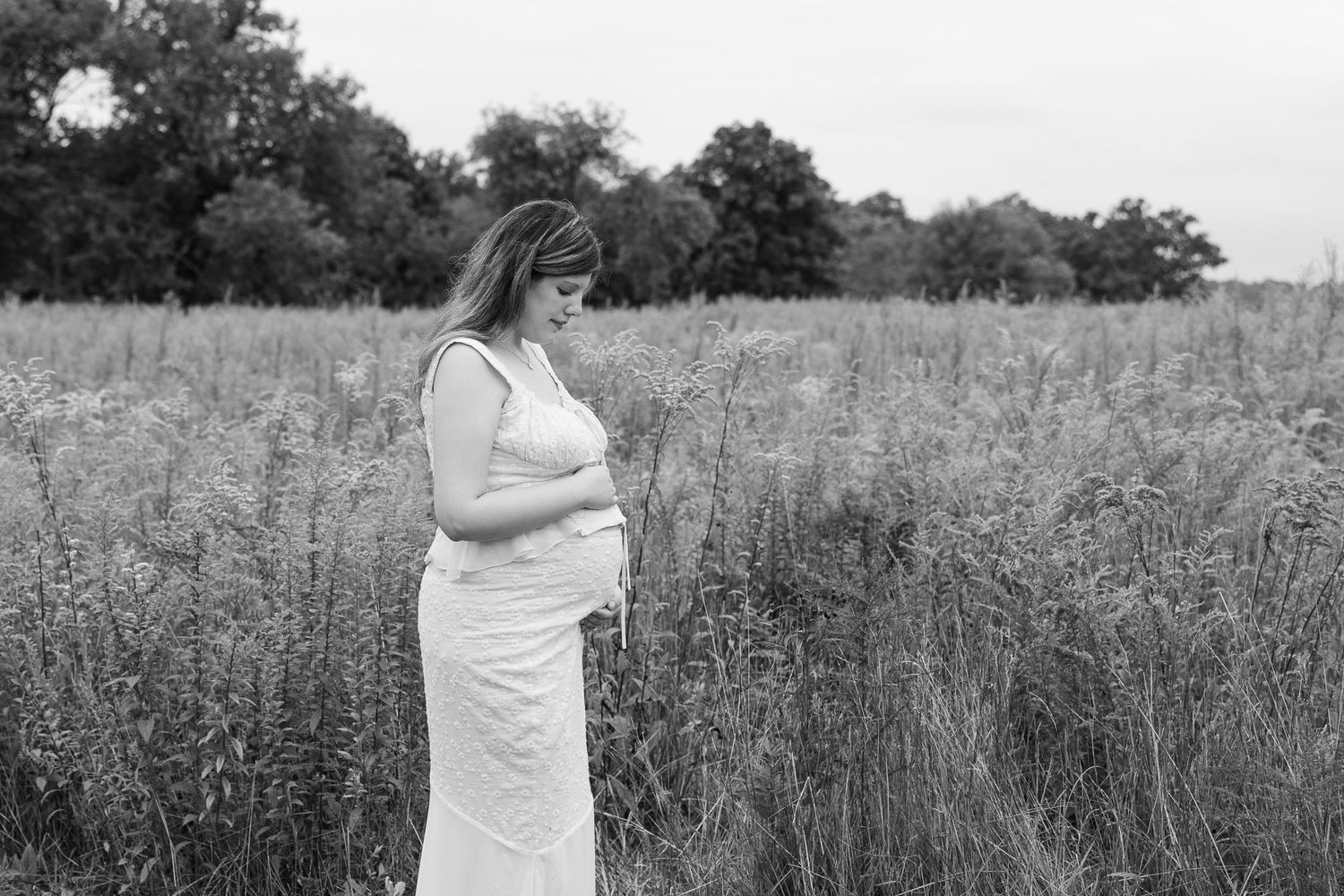 Monochrome side view of expectant mother in long dress amid tall grass.