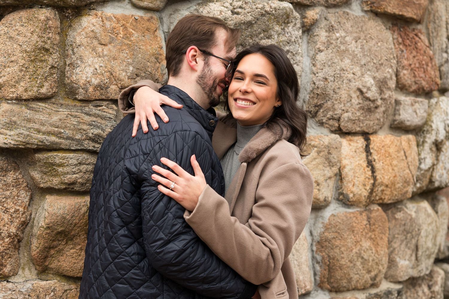 A couple embracing and smiling against a weathered stone wall background.