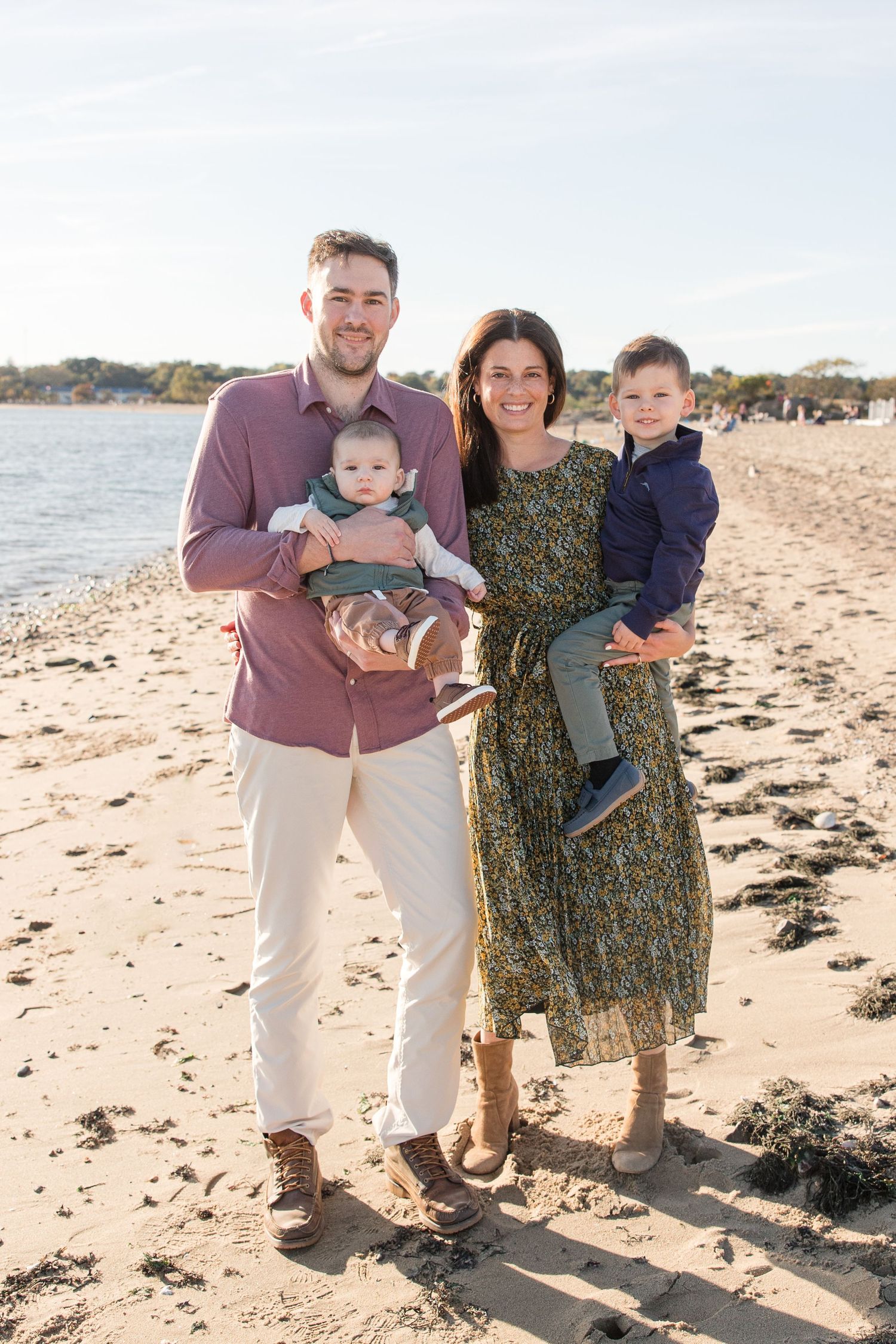 Family standing together on sandy beach with water and autumn trees in background.