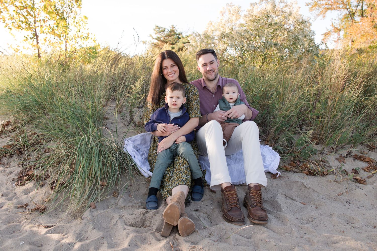 Family relaxing together on sandy dunes with tall grass in golden afternoon light.