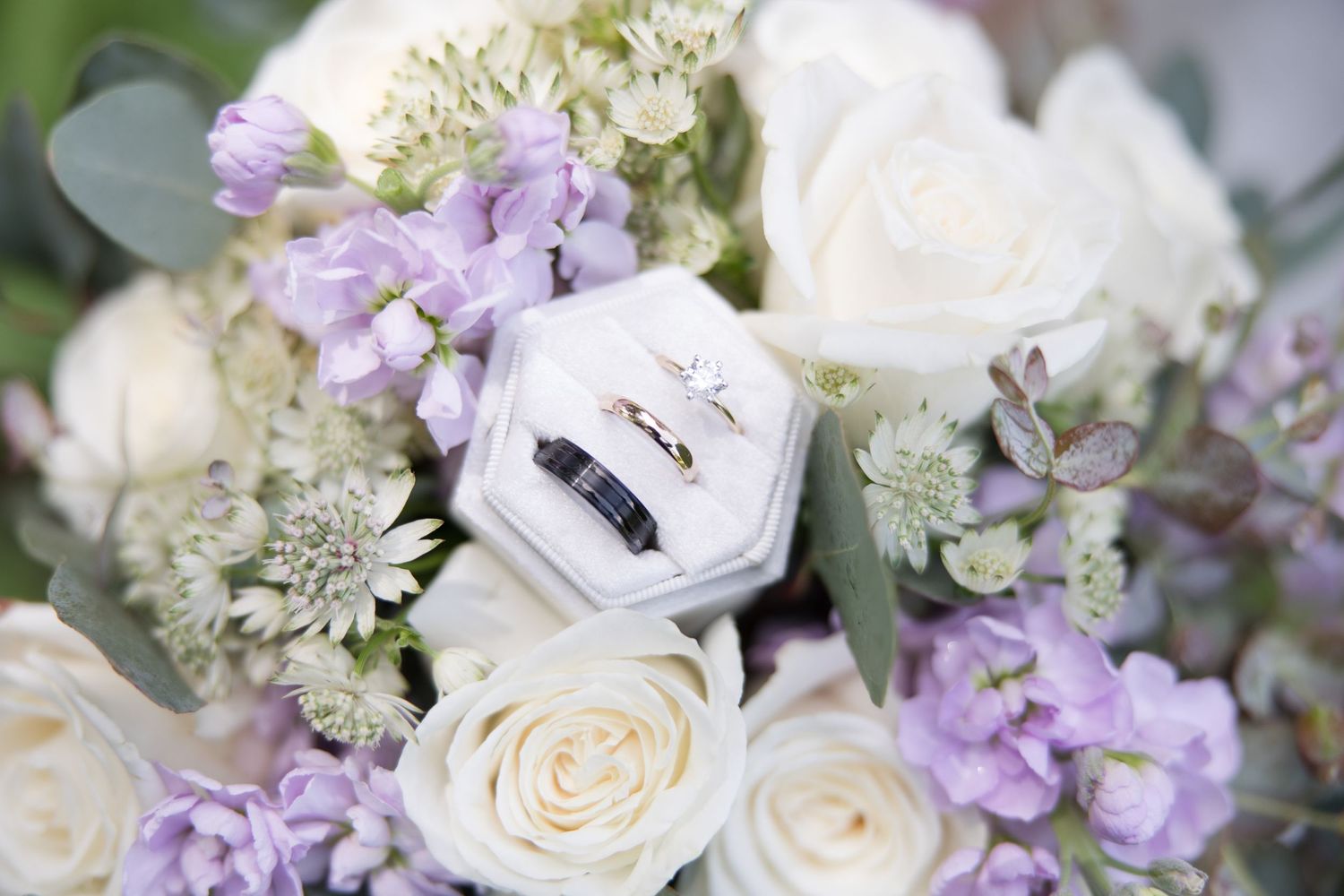 Wedding rings displayed in hexagonal box surrounded by white roses and purple flowers.