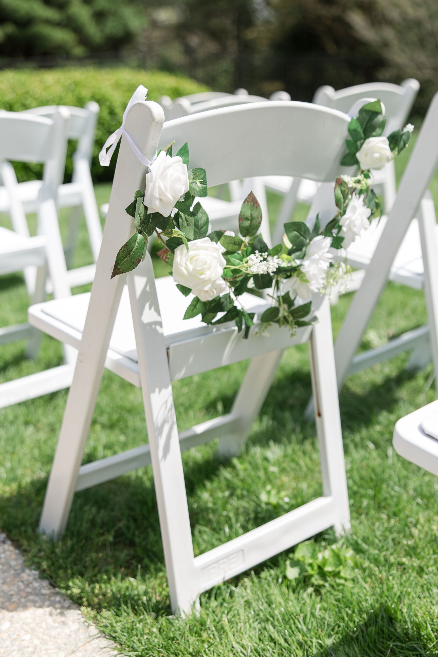 White folding chair decorated with fresh white roses and greenery garland for wedding ceremony.
