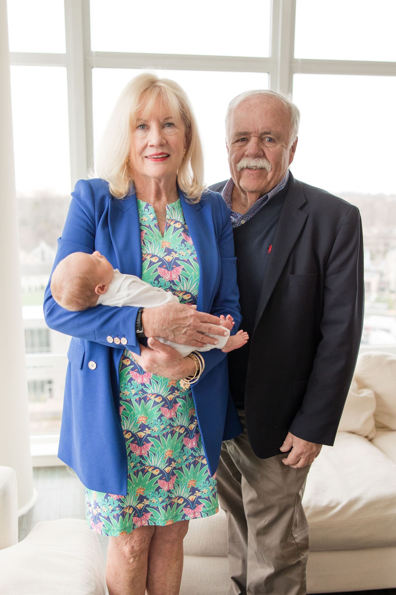 A couple in formal attire standing in a bright living room while holding a sleeping baby.