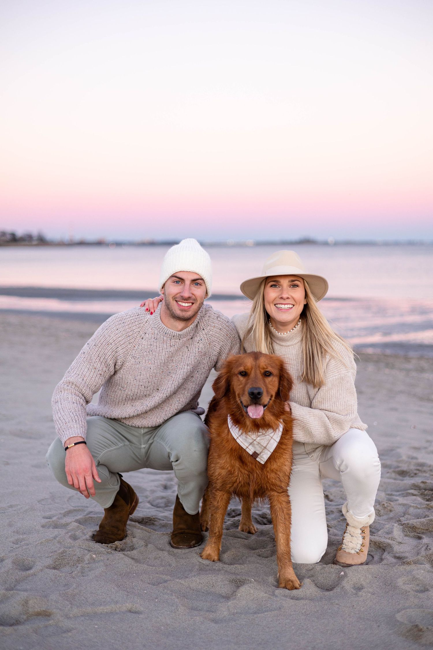 Golden retriever wearing bandana at beach with two people at sunset.